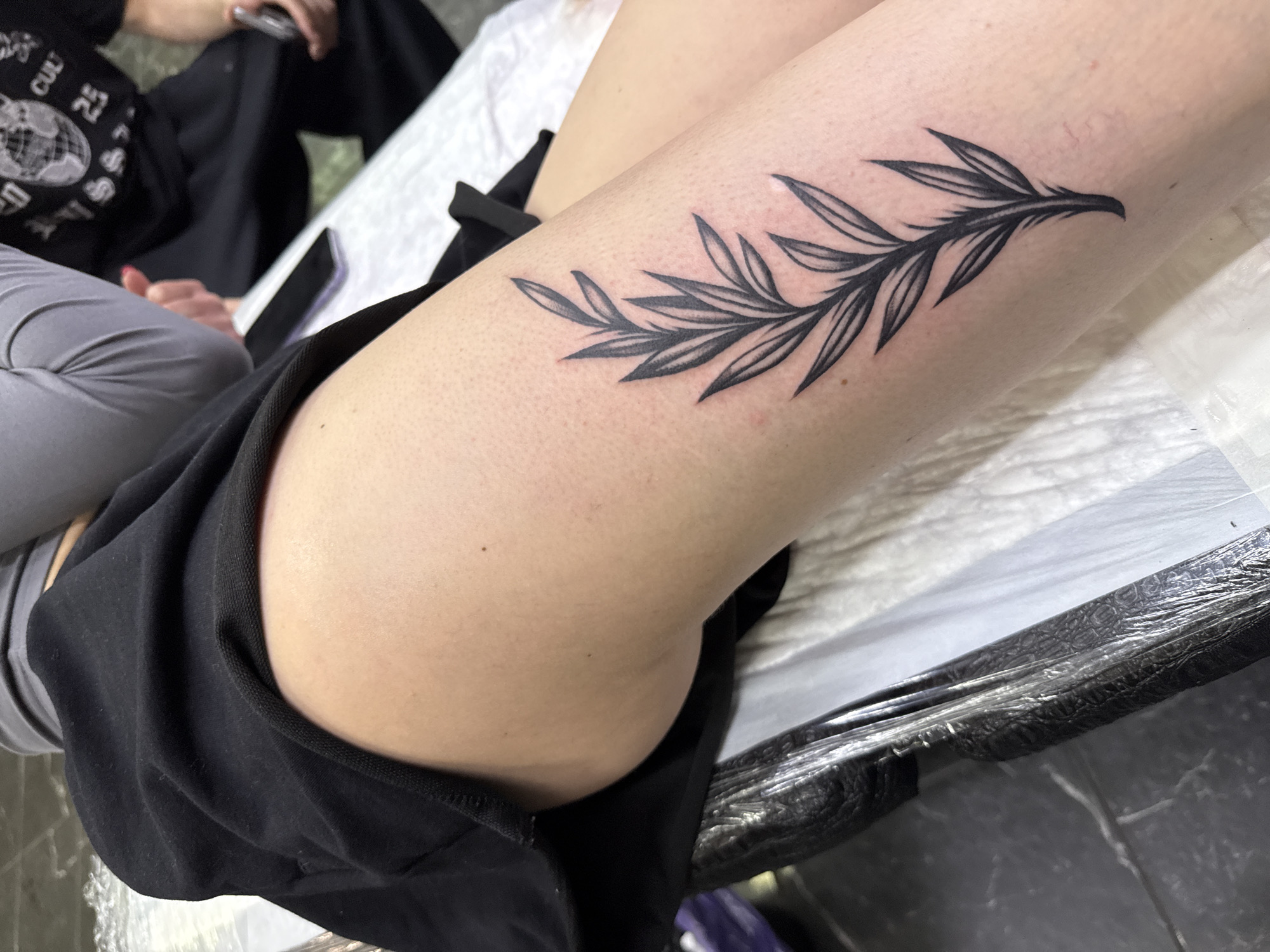 A close-up shot shows a tattoo of a stylized olive branch on a person's leg, presumably the calf or thigh. The tattoo is in black ink and appears fresh. The leg is resting on a surface covered with a white sheet. The individual is wearing black shorts, and the background reveals a glimpse of another person wearing grey sweatpants and a black shirt with a printed design. The surface beneath the white sheet is dark and possibly textured with some plastic lining.
There's no indication about the location of Moscow, Russia, in this image.