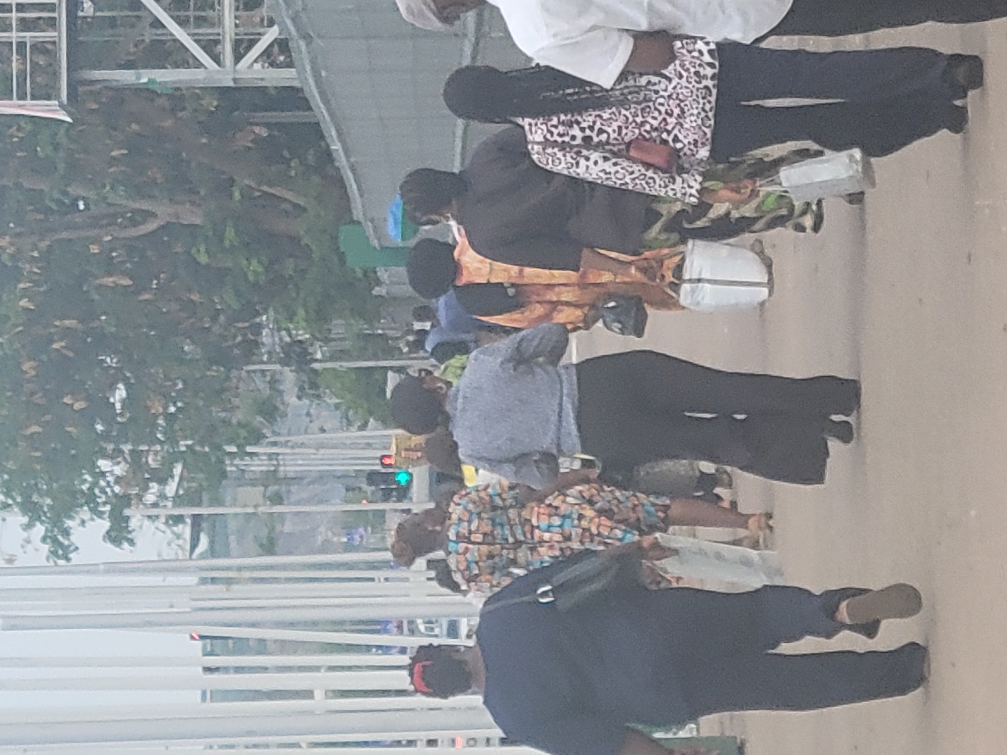 A group of people are standing on a sidewalk in Abuja, Nigeria. They are waiting to cross the street. There is a traffic light in the background that is green. The environment is urban, with trees and buildings visible.