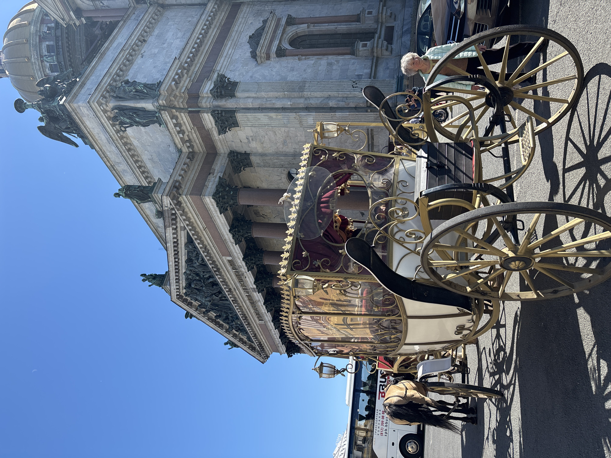 An ornate horse-drawn carriage, decorated with gold accents and crimson fabric, is parked on an asphalt surface in front of a grand stone building with a golden dome. The building, characterized by its classical architecture, columns, and sculpted figures, is Saint Isaac's Cathedral, a prominent landmark in Saint Petersburg, Russia. A light-colored bus is visible to the left of the carriage, and a brown car is parked to the right. An elderly woman, wearing a light blue jacket over a striped shirt and dark pants, stands near the carriage, looking towards the viewer. A horse, partially visible, is harnessed to the carriage. The sky is clear and blue, with sunlight casting distinct shadows on the ground.