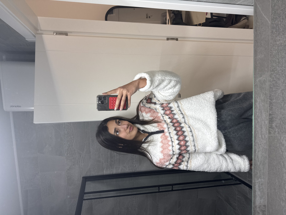 A young woman is captured in a mirror selfie, holding a smartphone with her right hand to take the picture. She has long, dark, straight hair that falls over her shoulders, dark eyes, and her lips are slightly parted in a neutral expression. Her fingernails are painted a light, neutral color.
She is wearing a cozy-looking, white, fluffy or fleece pullover sweater. The sweater features a decorative fair isle or Nordic-inspired pattern across the chest and upper arms, incorporating shades of light pink, white, and dark brown or grey. A black zippered or buttoned placket is visible at the neckline. She also appears to be wearing dark grey or charcoal-colored pants, possibly sweatpants or lounge pants.
The smartphone she holds is dark-colored, encased in a clear cover, and has a distinctive red accessory on its back, possibly a card holder or grip with multiple small, textured bumps.
The setting appears to be indoors, likely a bathroom or dressing area, as suggested by the large mirror and tiled walls. The wall behind her reflection (the actual background) is white, featuring what looks like a cabinet or door. Above this, an open storage area or shelf contains some dark items. To the left of her reflection, on the wall, a white rectangular device is mounted, possibly an air vent or appliance, with the text "dimola" visible on its lower edge. The lower half of the wall, visible in the foreground to her left and below her, is covered in grey, textured tiles, resembling stone or marble. A black frame, possibly belonging to a shower enclosure or room divider, is visible on the right side of the mirror's reflection. The overall lighting is even, suggesting indoor artificial light. The image is oriented sideways, appearing as if rotated 90 degrees counter-clockwise.