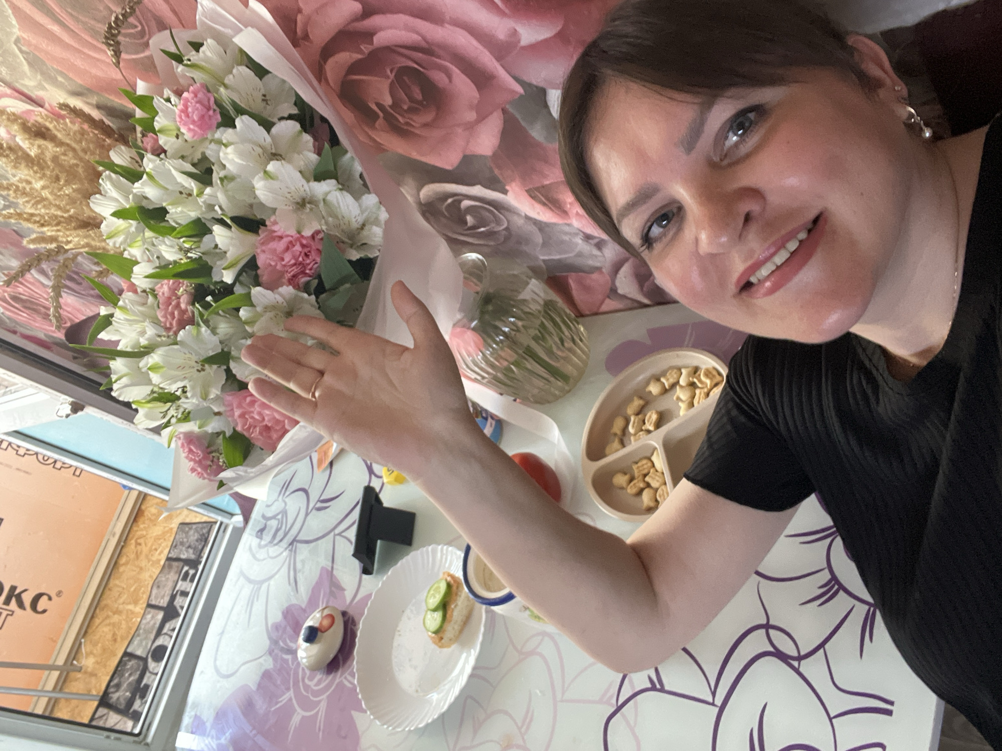 A medium shot captures a woman in a black top, lying on a table. Her arm is raised, with her hand waving towards the camera. A bouquet of white and pink flowers is situated behind her. The table is covered with a tablecloth featuring a floral design. On the table, there is a white plate with a piece of bread topped with cucumber slices, a small decorative item resembling a pastry, a small red object, a baby food plate filled with fish-shaped crackers, and a glass vase with water. In the background, a large bouquet is partially visible, and a floral patterned wallpaper lines the wall. The scene appears to be indoors, possibly a home.