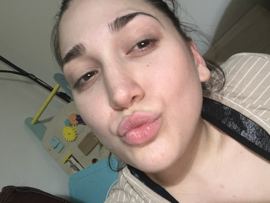 A fair-skinned woman puckers her lips towards the camera. Her eyebrows are dark and well-defined. She has brown eyes. She wears a tan and white striped shirt with a black trim. In the background, a child's interactive toy is visible with blue and yellow elements. Wires hang down the wall near the toy. The overall image has a casual, selfie-like quality.