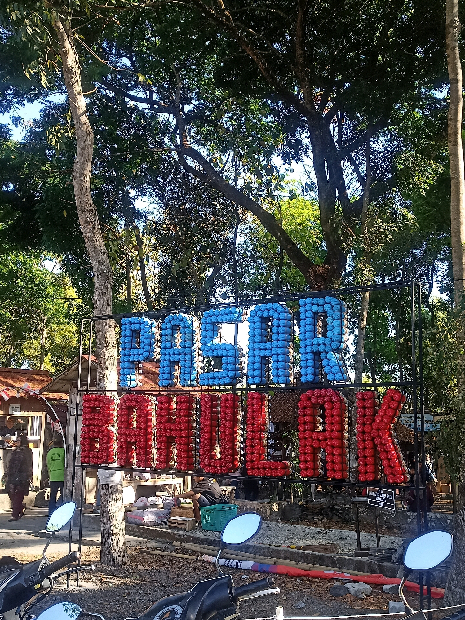 The image shows a sign with large, three-dimensional letters spelling "PASAR BULAK" in Indonesian. The letters are constructed from numerous small, brightly colored objects, with "PASAR" in blue and "BULAK" in red. The sign is mounted on a metal frame and is situated outdoors. In the background, there are trees with lush green foliage and a bright blue sky. Several structures, possibly stalls or small buildings with thatched roofs, are visible behind the sign. People are present in the scene, including individuals walking and standing near the stalls. The foreground shows parts of two motorcycles, suggesting a public area or roadside location. The overall environment appears to be a marketplace or a public gathering space in a tropical or subtropical setting. A small sign with text is visible on the right side of the image, and another sign is visible on the left behind the "P" of "PASAR".
