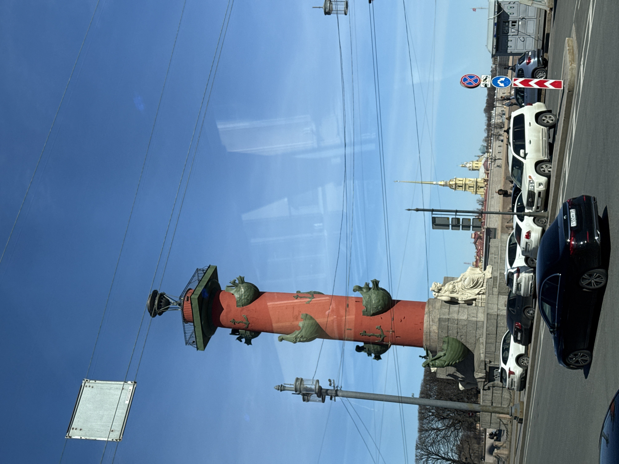 The image shows the Rostral Column in Saint Petersburg, Russia. The column is red with green bronze sculptural details. The Peter and Paul Fortress can be seen in the distance. The view is from a vehicle, evident by reflections in the glass and passing cars. There are no pedestrians visible in the scene. The weather appears clear and sunny.