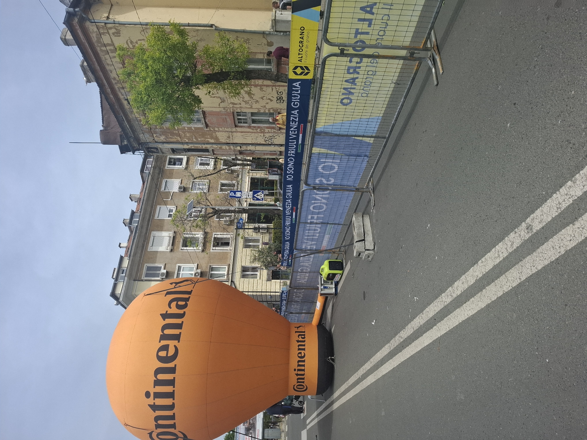 The image captures an urban scene in Burgas, Bulgaria, likely during a public event, featuring an orange "Continental" branded balloon prominently positioned on a street lined with buildings. The environment includes a security barrier with event-related signage ("FRIULI VENEZIA GIULIA IO SONO FRIULI VENEZIA GIULIA," and "ALTOGRANO Il cuore del grano"). A small electrical generator is visible near the balloon. The street has white double lines, indicating traffic lanes. The architecture comprises aging buildings with windows and trees. The composition suggests the photo was taken during the daytime, under relatively clear skies. Pedestrians are partially visible near the balloon.