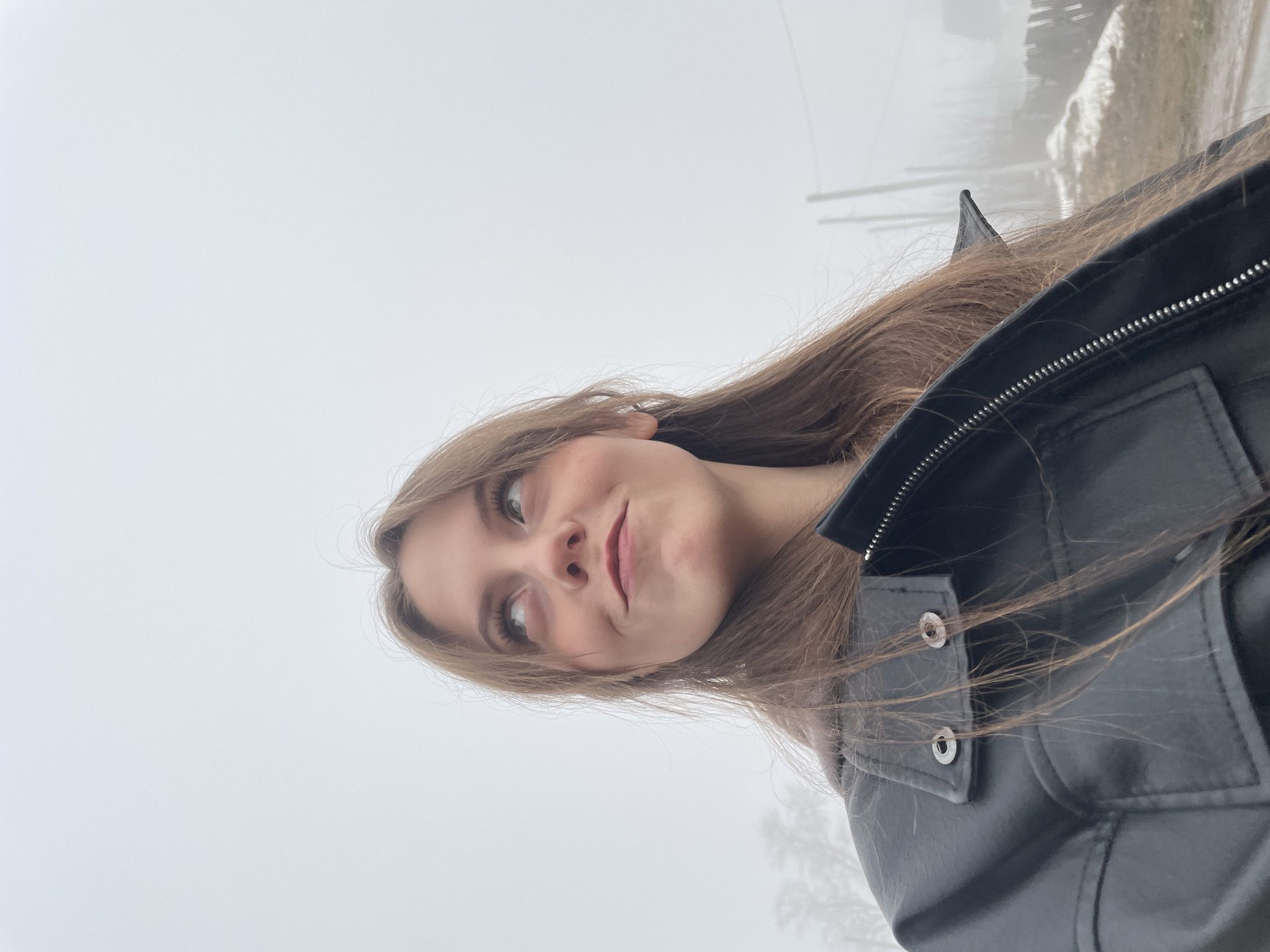 A young woman is standing in a foggy, outdoor location. She is wearing a black leather jacket. Her brown hair is long and loose. She is looking off to the side with a slight smile. The background appears to be a street with poles, possibly in a rural or suburban area, obscured by heavy fog. While the location is alleged to be Фоки, Russia, there is nothing in the image to independently verify this claim.