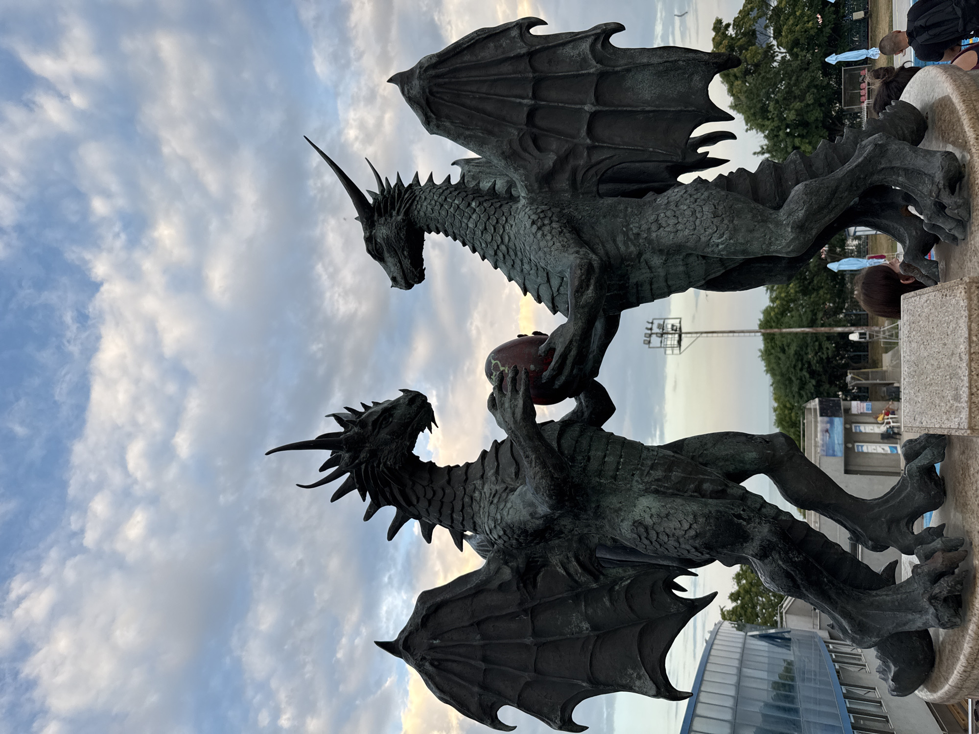 The image shows a sculpture of two bronze dragons with outstretched wings, seemingly in mid-air. The dragons are dark, with detailed scales and sharp claws. One dragon is positioned above the other, with its claws holding a reddish-brown orb, possibly representing a treasure or a mythical artifact. The dragons' heads are turned towards each other, with their mouths open in what could be a roar or a greeting.
The background consists of a cloudy sky with hints of sunset light breaking through, suggesting it is either early morning or late evening. Trees and a modern building with reflective windows are visible behind the sculpture. In the distance, there are also hints of people and structures, possibly indicating a public space or park.
The overall mood of the sculpture is powerful and dramatic, conveying a sense of fantasy and myth. The lighting and the cloudy sky add to the atmospheric quality of the scene. No visible text is present in the image. The sculpture appears to be a notable landmark, possibly in Varna, Bulgaria, given the location context.