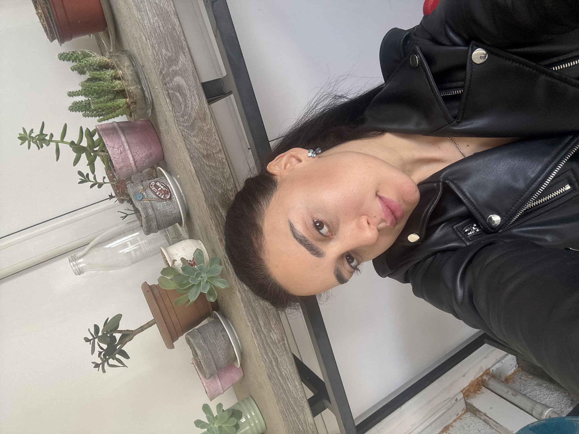 A woman in a black leather jacket stands indoors near a shelf of potted plants, including succulents and cacti. A small glass bottle is also on the shelf. The location is presumed to be Tbilisi, Georgia as that was the source of the prompt.