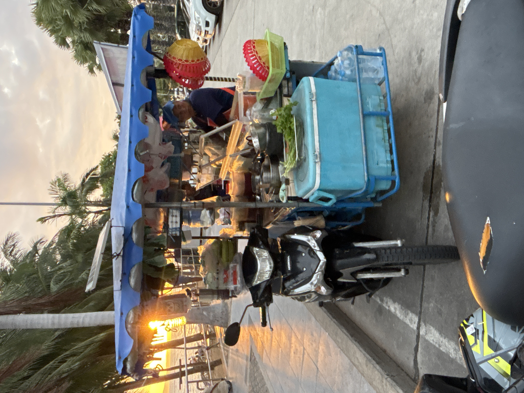 The image depicts a street food cart at sunset in Pattaya City, Thailand. The cart, which is blue and white, is stocked with various food items, cooking utensils, and a turquoise cooler. A man wearing a blue cap and an orange apron stands behind the cart, appearing to serve customers. A black motorcycle is parked in front of the cart, and another motorcycle is partially visible in the foreground. The background shows palm trees, a beach with people, and the ocean, all bathed in the warm light of the setting sun. Several cars are parked in the background. The scene captures a typical evening street food vendor setting in a coastal city.