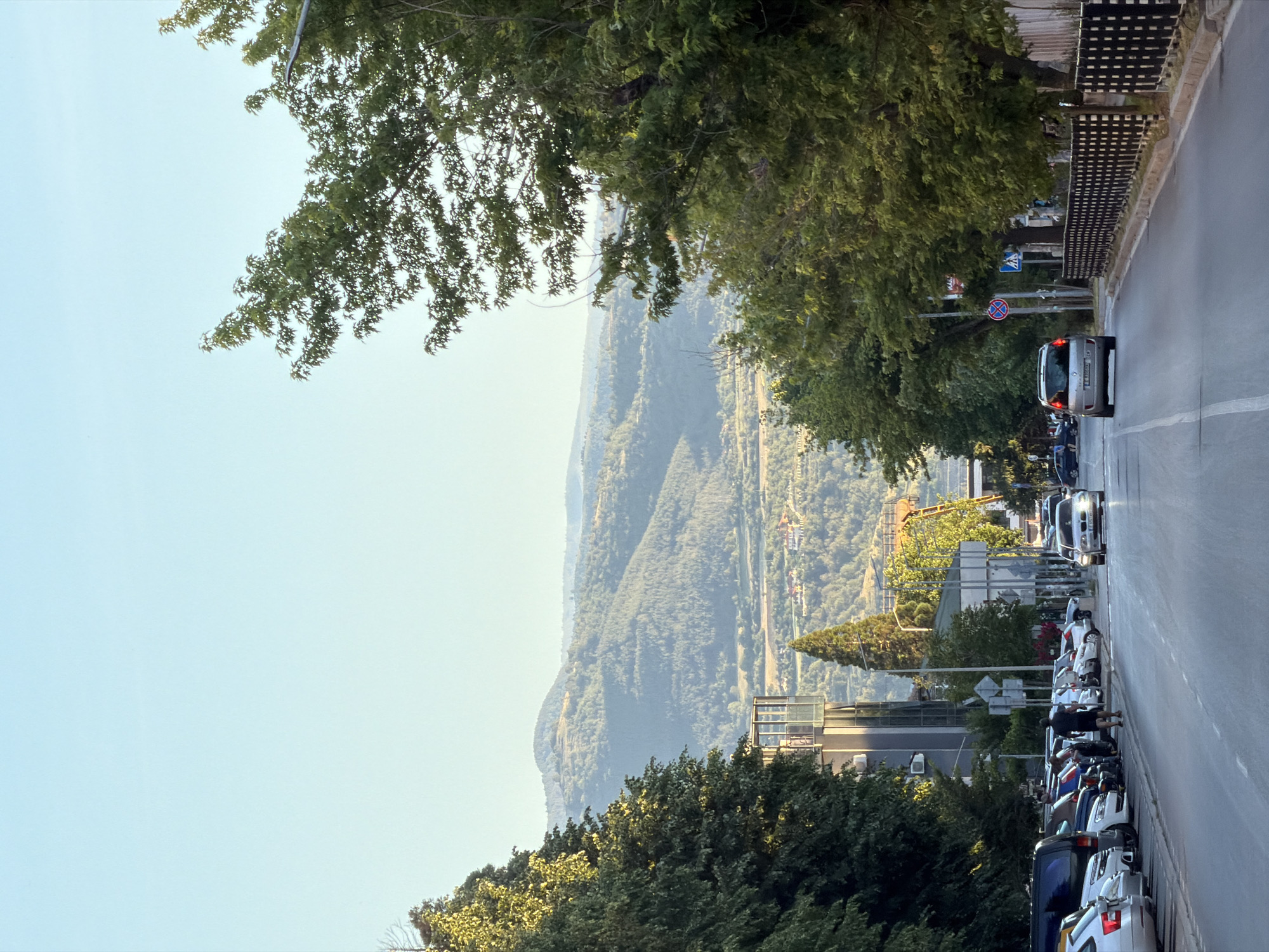A scenic view in Blagoevgrad, Bulgaria, features a street lined with parked cars and lush trees, overlooking a valley with rolling hills. A silver car drives on the right side of the road. The backdrop includes a vista of mountains shrouded in a slight haze. The sky is clear and light blue. Street signs are visible along the roadside. Buildings are interspersed among the trees. The scene appears to be captured on a sunny day.