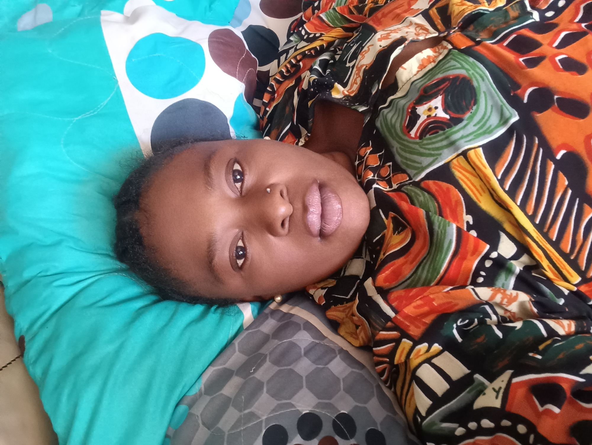 A person with dark skin and short dark hair is lying on a bed with a patterned bedspread. The bedspread has sections in turquoise with white and blue circles, gray with dark gray honeycomb pattern, and tan. The person is wearing a colorful patterned garment of black, orange, white, green, and yellow. The person has a nose piercing on the left nostril, light-colored lips, and is looking at the camera. The location cannot be verified as Jos, Nigeria from the available data.