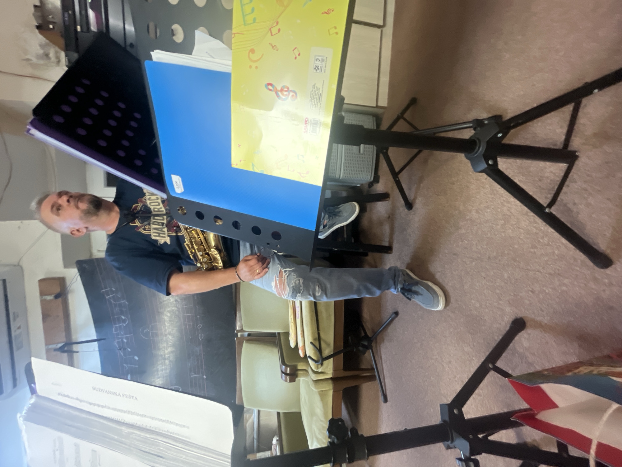 The image depicts an indoor scene with a man holding a saxophone, seemingly in a rehearsal or performance space. The man, with short, grey hair and a beard, wears a dark t-shirt with the text "HARD ROCK" and distressed blue jeans. He is seated and looking slightly to his left. Several music stands are visible, holding sheet music and folders. One prominent stand features a bright blue folder and a yellow folder with musical notes printed on it. In the background, two green chairs are visible, and further back, what appears to be a blackboard with musical notation. The floor is carpeted in a muted tone. The overall impression is that of a casual music practice or studio environment. The provided context indicates the location is in Budva, Montenegro.