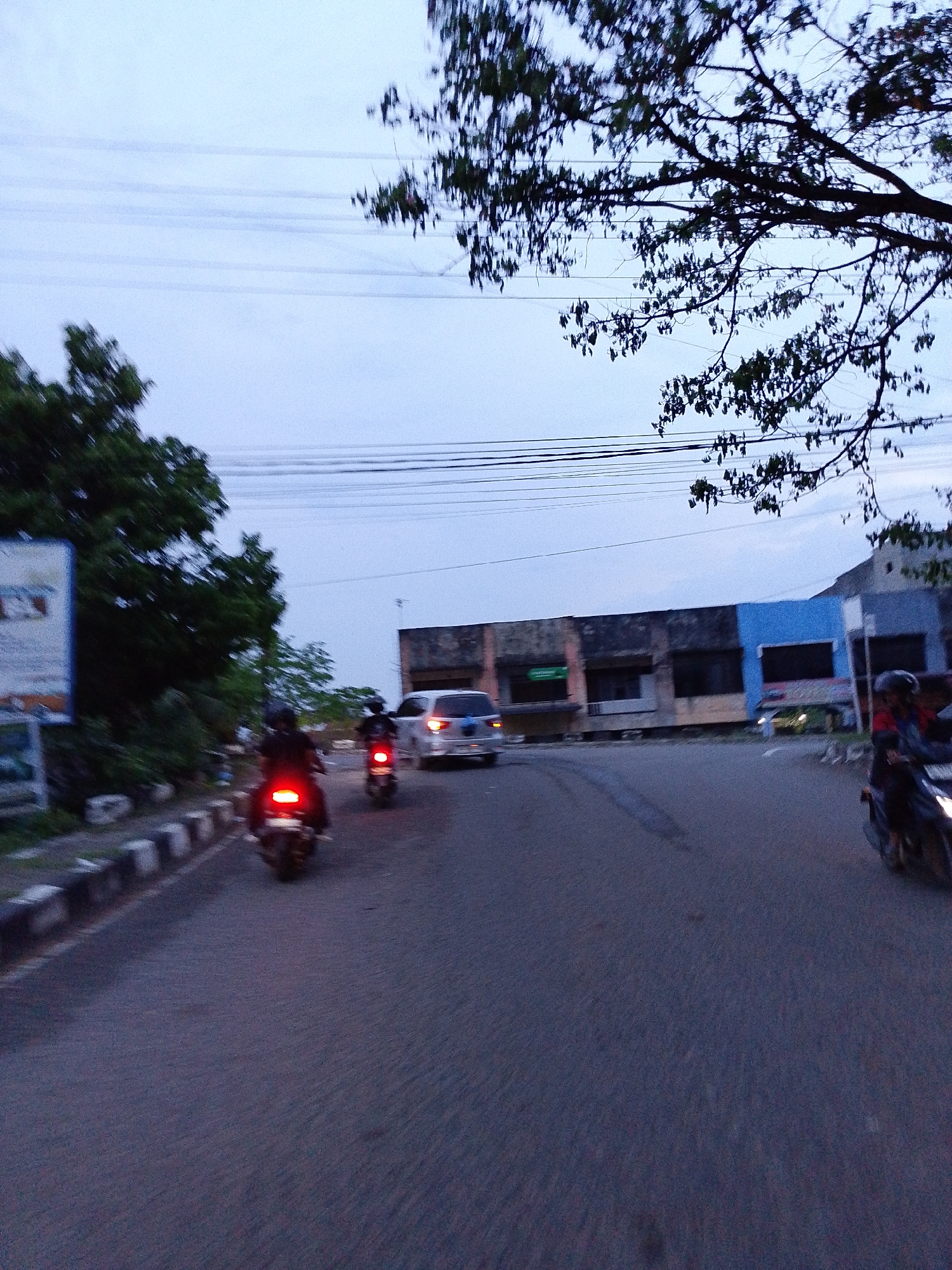 This image captures a street scene in Lhokseumawe, Indonesia, characterized by dim, diffused lighting, suggesting either dusk, dawn, or an overcast day. The sky is a muted, hazy blue-grey, with several horizontal power lines traversing it.
In the foreground, an asphalt road dominates the lower half of the frame. At least three motorcycles are visible, with two prominently displaying their illuminated red taillights, indicating active travel or braking in low light. The riders are wearing helmets. One motorcycle is centrally positioned, moving away from the viewer, while another is further ahead on the right. On the left side of the road, a sidewalk is delineated by a low curb painted with black and white stripes.
The midground features a line of buildings on the right side of the street. These structures display varied facades, incorporating colors such as blue, brown, and grey, and appear somewhat aged. A white car is visible among the traffic, positioned closer to these buildings. On the left side of the street, a large, rectangular billboard or sign stands partially within the frame, but any text on it is illegible due to blur and distance. Dense green foliage and tree branches, some silhouetted against the sky, frame the scene from the left and upper right corners. The activity primarily involves vehicles in motion on the street. No discernible emotions are visible, and the scene depicts typical daily life in an urban setting.