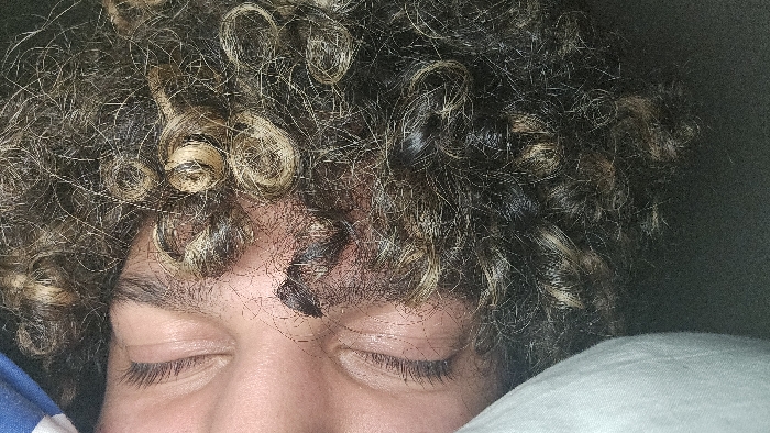 A close-up captures a person with closed eyes, suggesting sleep or relaxation. Their curly hair, a mix of blonde and darker shades, dominates the upper portion of the frame. Part of their face, including the nose and eyelids, is visible. The person rests on a white pillow and is partly covered by a blue and white striped fabric on the left. The background fades into a dark, indeterminate space.