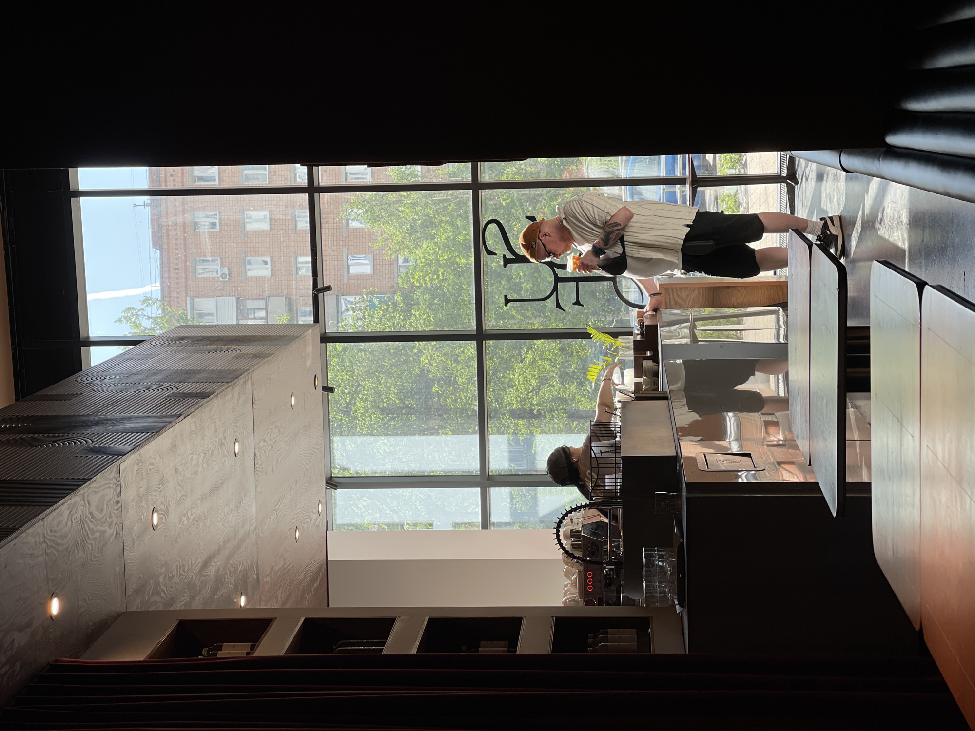 The image shows the interior of a cafe or restaurant in Izhevsk, Russia. A person wearing a light-colored striped shirt, dark shorts, a brown cap, and sneakers is standing at a counter, holding a cup and looking down at it. The counter is in front of a large window with a view of a brick building and green trees. The cafe's logo is visible in the window. Below the counter is an espresso machine and various glasses. The interior features wooden walls, recessed lighting, and dark curtains or upholstered benches. A staircase with red carpet is visible in the lower left. The image is taken from a low angle, looking up toward the window.