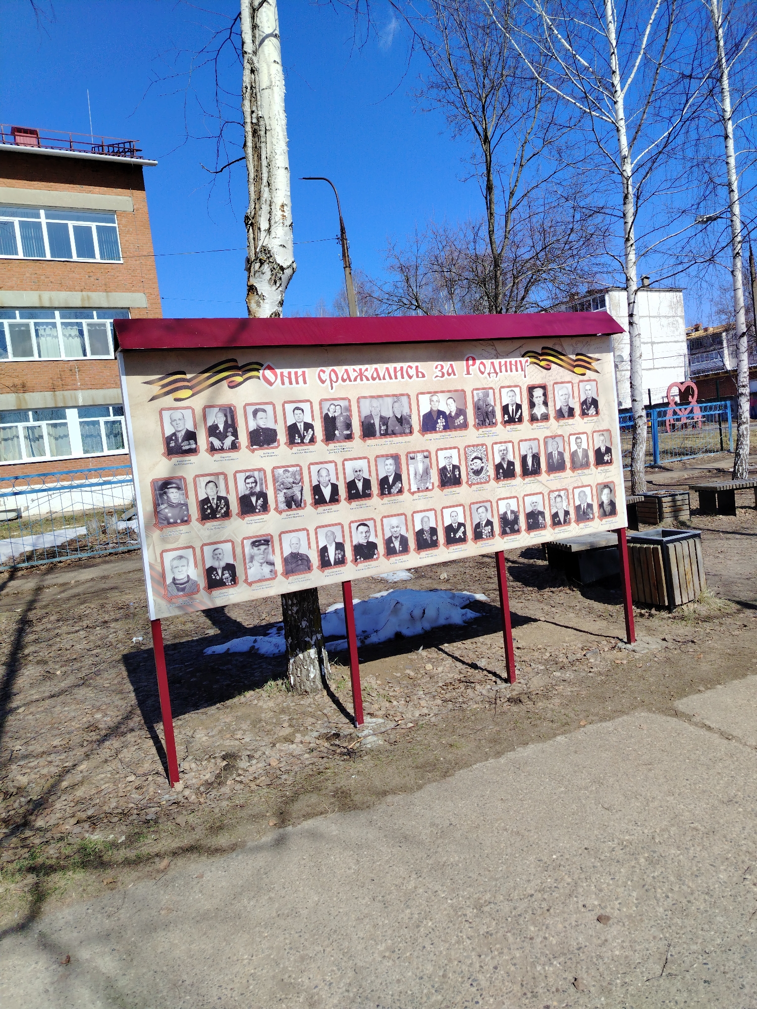 The image shows a memorial stand in Noviy, Russia, featuring portraits of veterans. The stand has a red border and the inscription "They fought for the Motherland" in Russian. It is positioned in a public outdoor space with trees, benches, and a playground visible in the background. A brick building stands nearby, under a clear blue sky. Patches of snow remain on the ground.