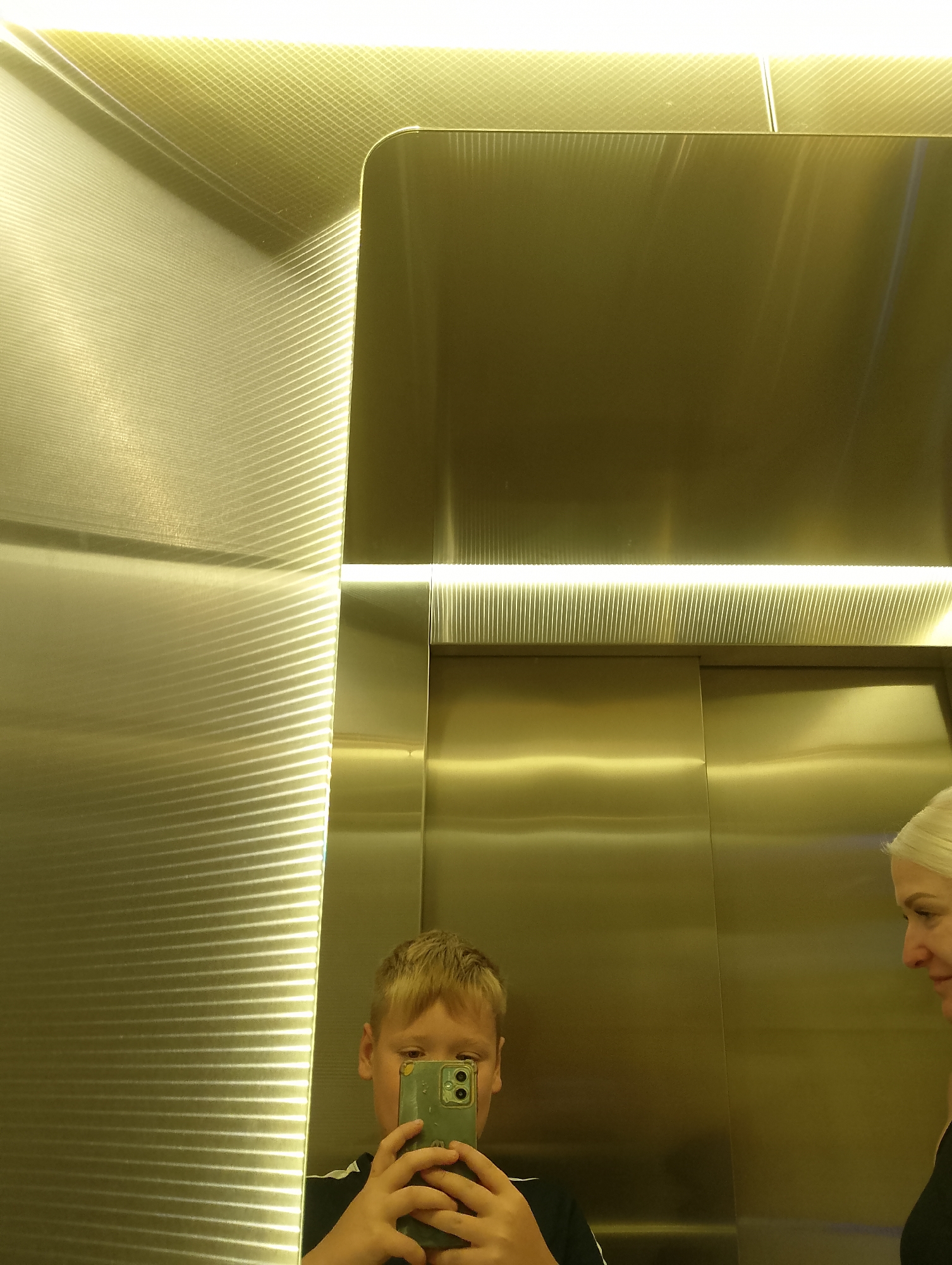 A young boy with blonde hair is taking a mirror selfie in an elevator. He is holding a smartphone with a green case and is wearing a black t-shirt with white stripes. To the right of the boy, a woman with blonde hair is visible in the reflection, looking at the boy. The elevator's interior is covered in reflective gold-toned metal panels, with a horizontal light fixture running along the top of the elevator doors and another light running vertically along the left wall. The lighting creates a bright and somewhat distorted reflection. The scene appears to be an elevator in Chaiky, Ukraine.