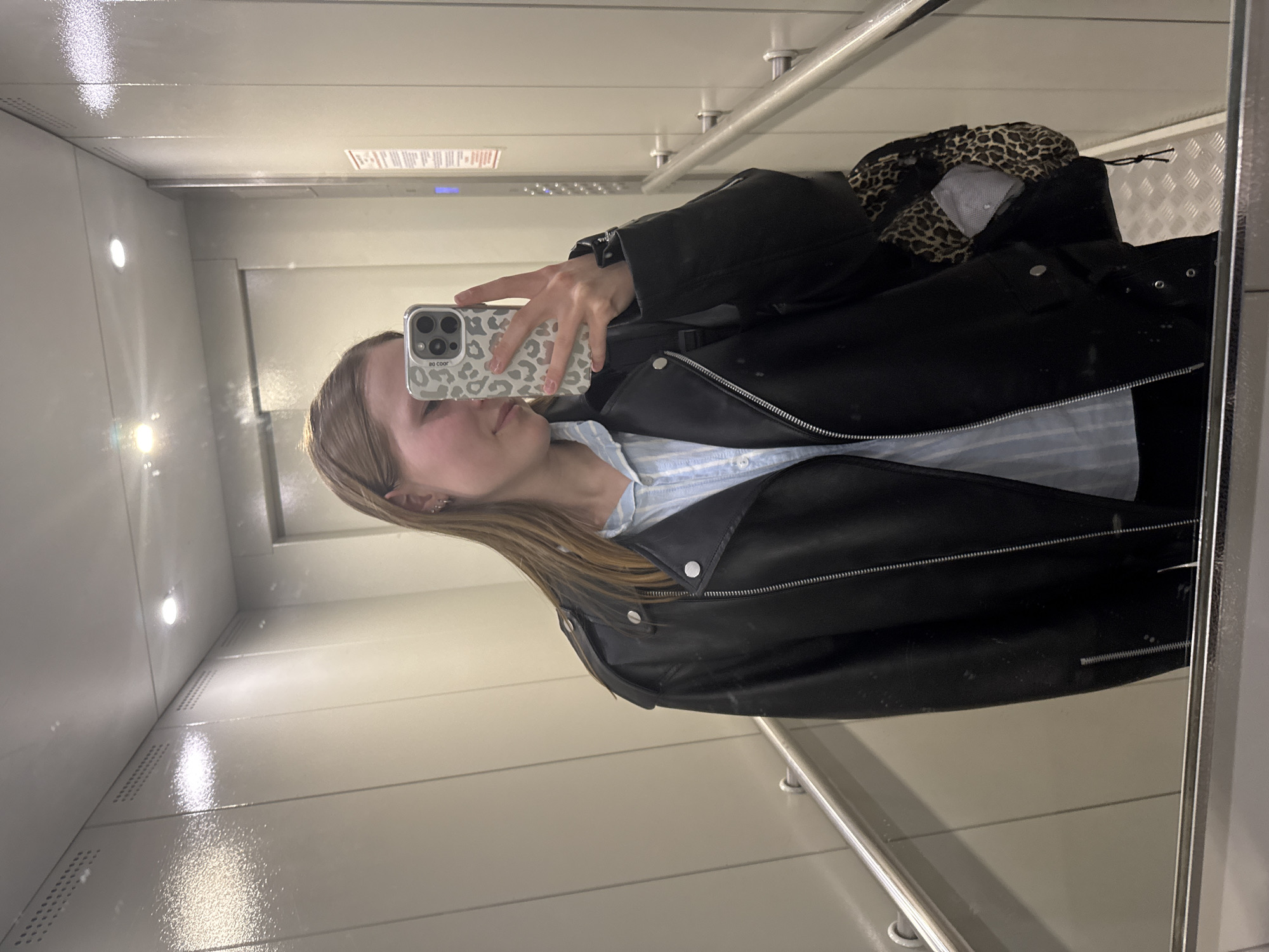 The image depicts a young woman taking a selfie in an elevator. She is holding a phone with a patterned case. She is wearing a black leather jacket over a light blue striped shirt. A leopard-print backpack is visible behind her on the right. The interior of the elevator is white, with metal bars and a mirror on the right. The lighting is bright and diffused. The location of the city, Chaikovsky, Russia is unidentifiable.