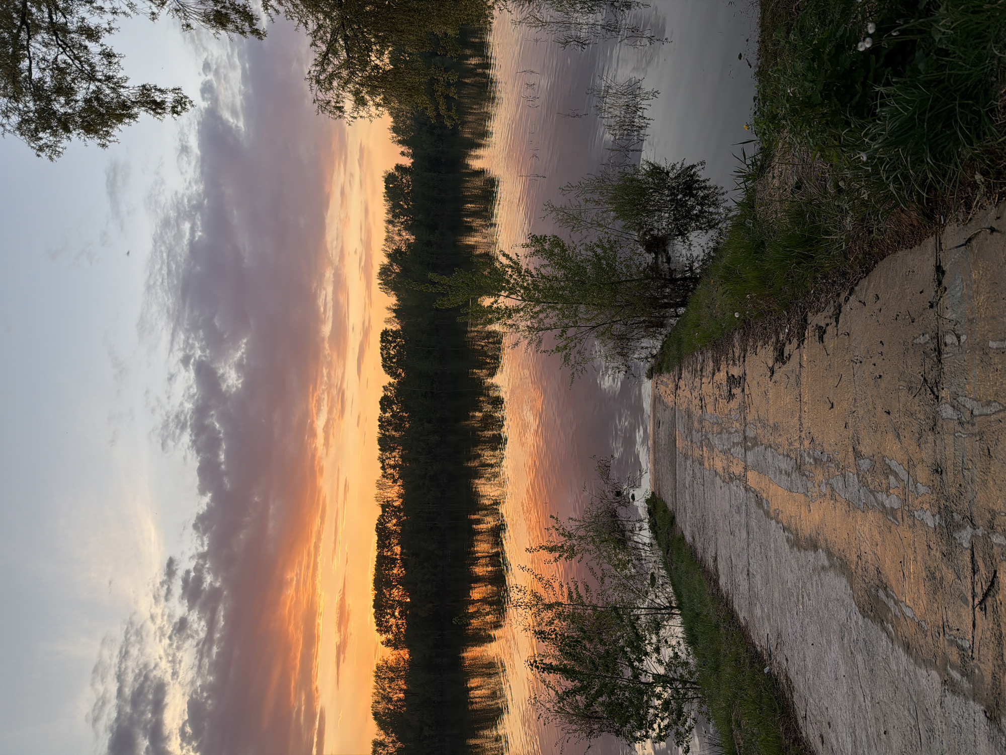 The image captures a sunset scene at a location with water and a path. The sky displays a gradient of colors, from light gray to orange, reflecting in the water. Trees line the bank opposite the path, mirrored in the water. The path is concrete or stone and is lined with grass and some shrubbery. The location appears to be in Chaikovsky, Russia. There are no visible people or interactions.