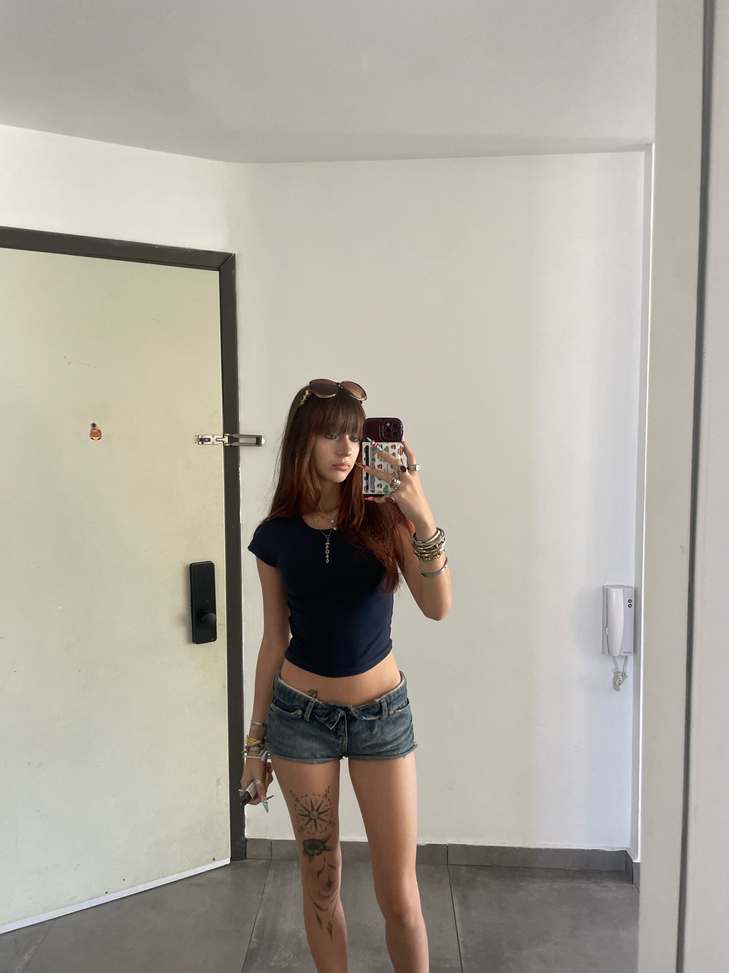 A young woman with long, dark brown hair and bangs, wearing sunglasses on her head, is taking a mirror selfie. She is dressed in a dark blue cropped t-shirt and distressed denim shorts. She is adorned with multiple bracelets and rings, and has a necklace with a star pendant. She has a tattoo on her left thigh that appears to be a compass rose with other designs. The setting is indoors, with a white wall and a large mirror reflecting the woman. To the left of the woman is a light-colored door with a black door handle and a silver latch. To the right of the woman, on the white wall, is a white intercom device. The floor is tiled in a grey pattern. The overall environment suggests a modern interior, possibly a hallway or dressing area within a home or hotel. The image does not contain any specific visual cues to indicate a location in Tel Aviv, Israel.