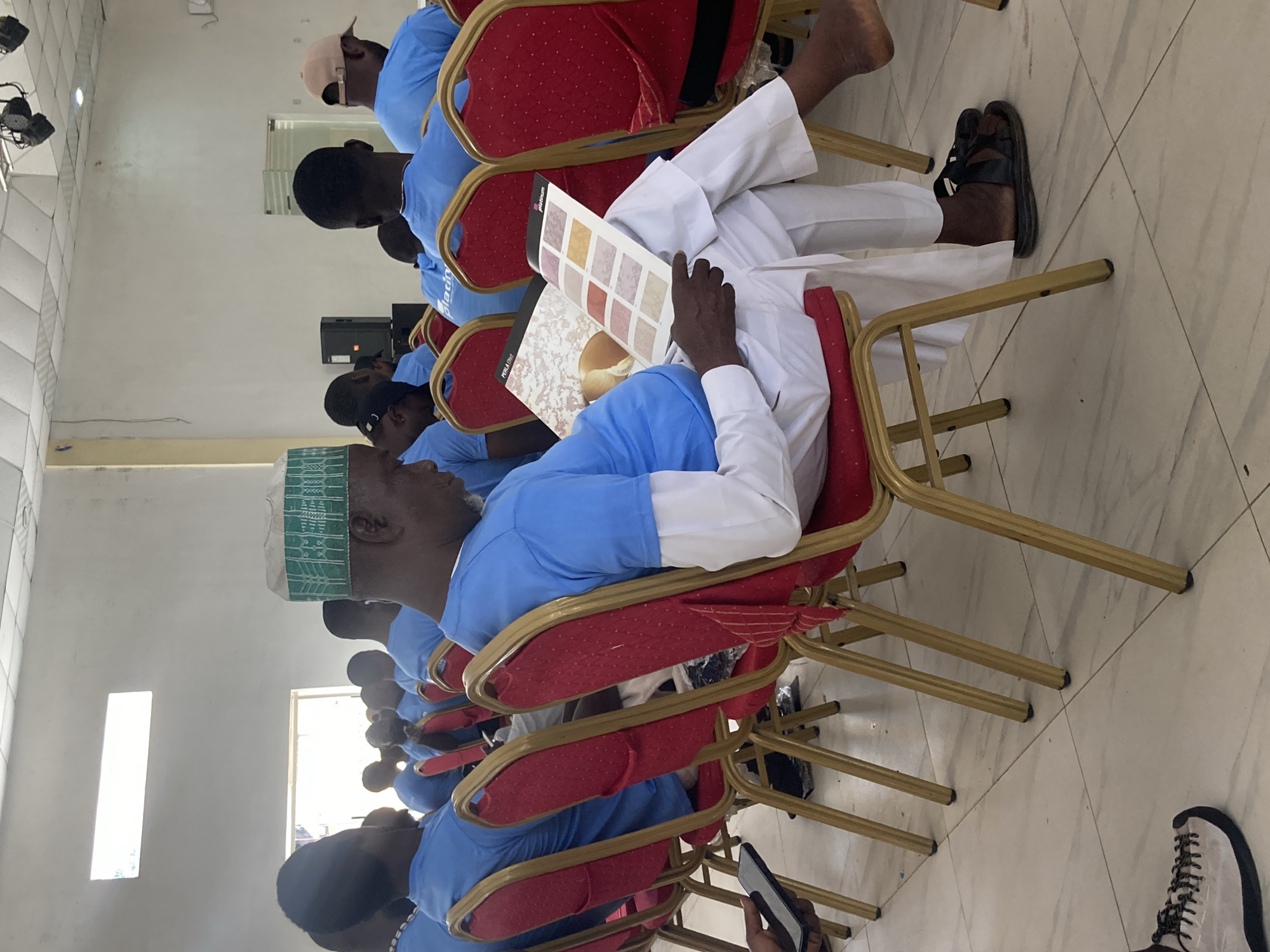 A group of men are seated in a row of red chairs, seemingly in a conference or training setting. The men are wearing light blue shirts, and some are wearing white caps. One man in the middle is looking at a book or a binder containing swatches of paint colors. The men are oriented towards the front of the room, likely listening to a speaker or presentation. The setting appears to be an indoor room with light-colored tiled floors and white walls. The image was taken at a location in Jos, Nigeria.