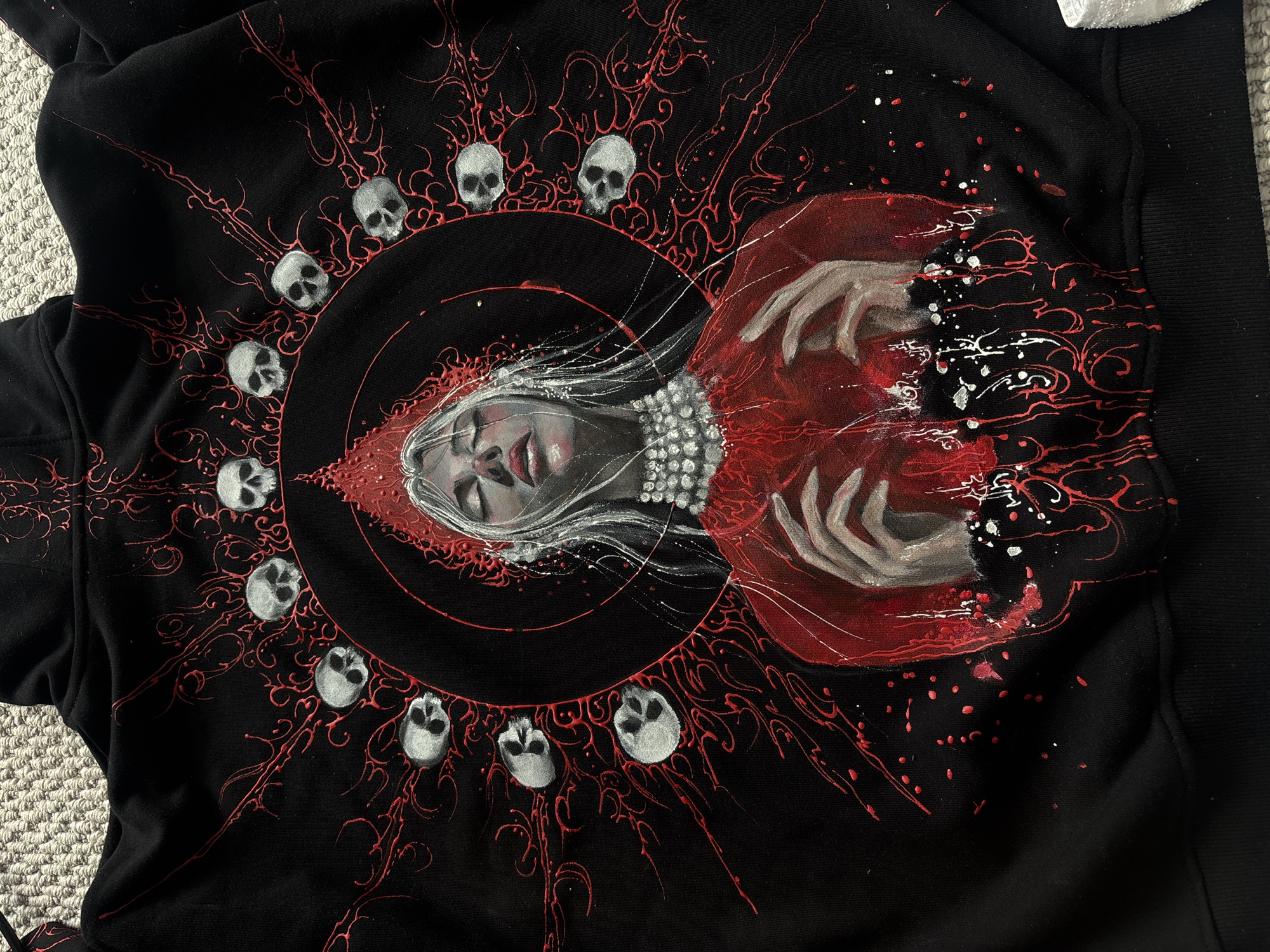 The media file displays a custom-painted black garment, likely a hoodie or sweatshirt, adorned with a dark, gothic, and mystical artwork.
**Subjects:** The central subject is a stylized female figure, possibly a witch, vampire, or ethereal being. She has pale skin, prominent red lips, closed or heavily shadowed eyes, and long, flowing white hair that cascades around her face. She wears an elaborate, beaded white necklace or collar and a voluminous red garment or cloak, which billows around her with visible brushstrokes and white paint splatters. Her hands are depicted in a dramatic gesture, reaching out or drawing inward.
**Objects:** Surrounding the central figure is a circular arrangement of approximately twelve small, grey or silver-colored skulls, each detailed with eye sockets and nasal cavities, creating a halo or crown effect.
**Scene/Setting:** The artwork is set against a stark black fabric background. Intricate, swirling patterns in red paint emanate from behind the central figure and the circle of skulls, spreading across the garment like vines, blood, or energy currents. These red patterns are dynamic, with additional red and white paint splatters giving the impression of movement or an abstract cosmic effect.
**Activity or event taking place:** The image captures a detailed piece of wearable art, likely depicting a supernatural or fantasy scene. The figure's expression and posture, combined with the skull motifs and swirling red elements, evoke a sense of drama, power, or melancholy.
**Notable details:** The use of contrasting colors—deep black, vibrant red, and stark white/silver—enhances the artwork's impact. The texture of the paint is visible, particularly in the red swirls and white splatters, suggesting a hand-painted quality. The ribbed cuff of a sleeve is visible on the right, confirming the garment type.
**Visible text:** There is no visible text in the image.