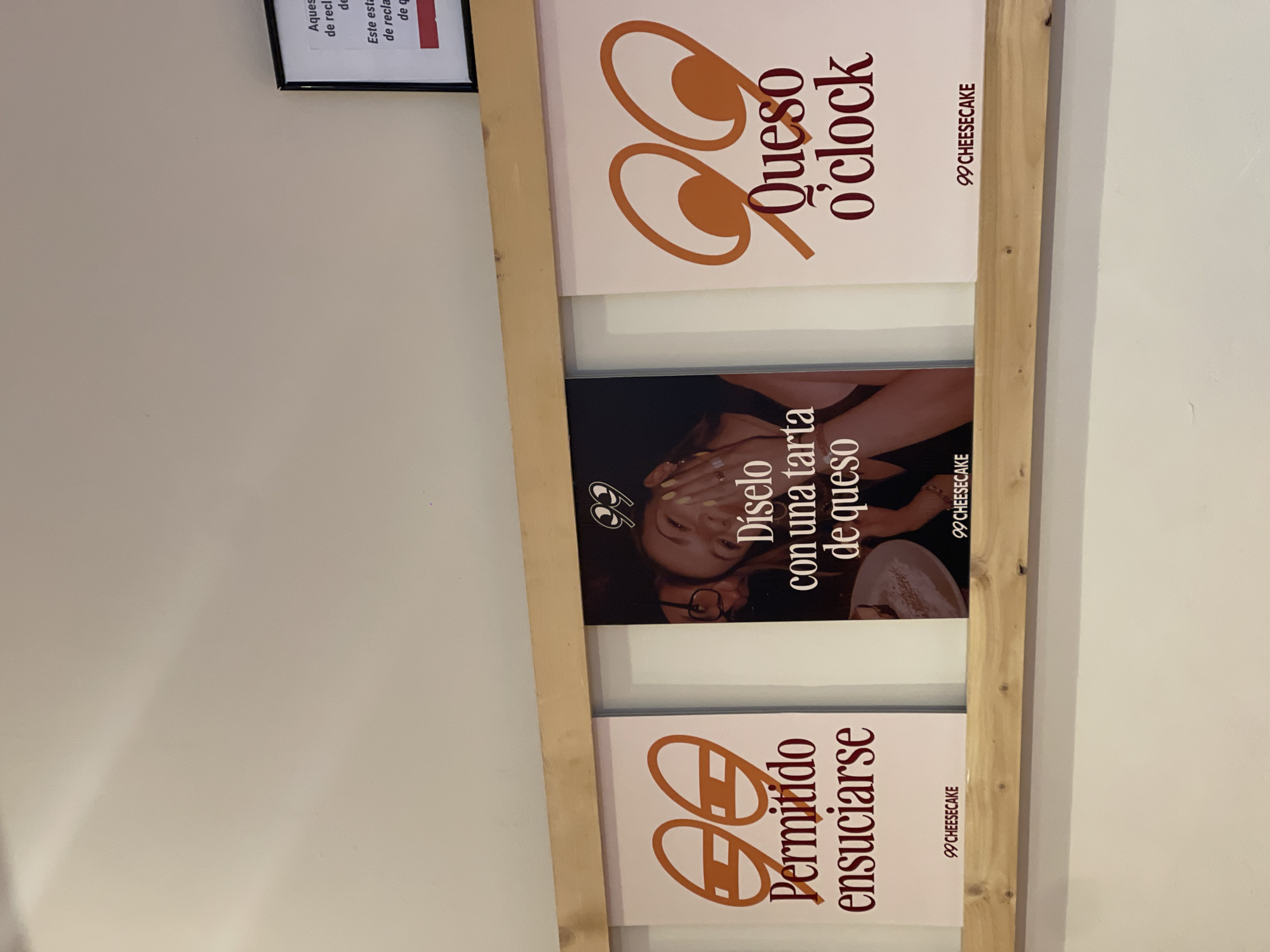A vertical, medium-shot photograph depicts a wooden frame displaying three promotional posters for the "99 Cheesecake" brand. The posters are arranged vertically, one above the other, against an off-white wall. The top poster reads "Queso o'clock" in a modern font with stylized eye-like shapes. The middle poster features a woman with cheesecake on her face and the text "Díselo con una tarta de queso," meaning "Say it with a cheesecake". The bottom poster reads "Permitido ensuciarse," or "Allowed to get dirty" also with stylized eye-like shapes. The brand logo, "99 Cheesecake," is visible in the bottom right corner of each poster. A framed sign with partially visible text is mounted above and to the left of the wooden frame. Given the Spanish text on the posters, the location is likely in València, Spain.