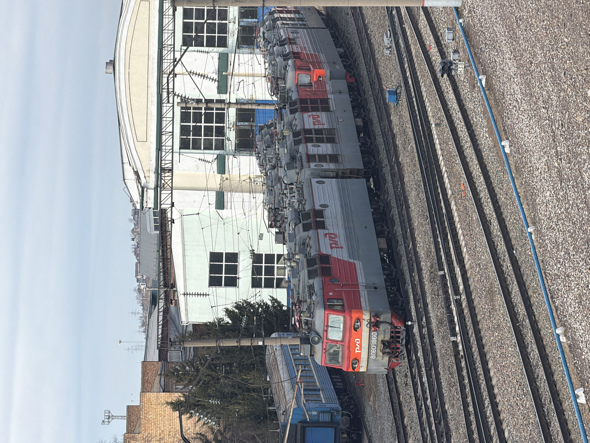 A train yard in Krasnoyarsk, Russia, features several gray and red Russian Railways (RZD) locomotives on multiple tracks. An RZD electric locomotive, marked "ВЛ80-4860," is at the forefront. An adjacent blue train car is also present. A large, light-colored industrial building with multi-paned windows is in the background. The ground is covered in gravel. Overhead power lines and support structures are visible, along with various utility poles and cables. The sky is overcast.