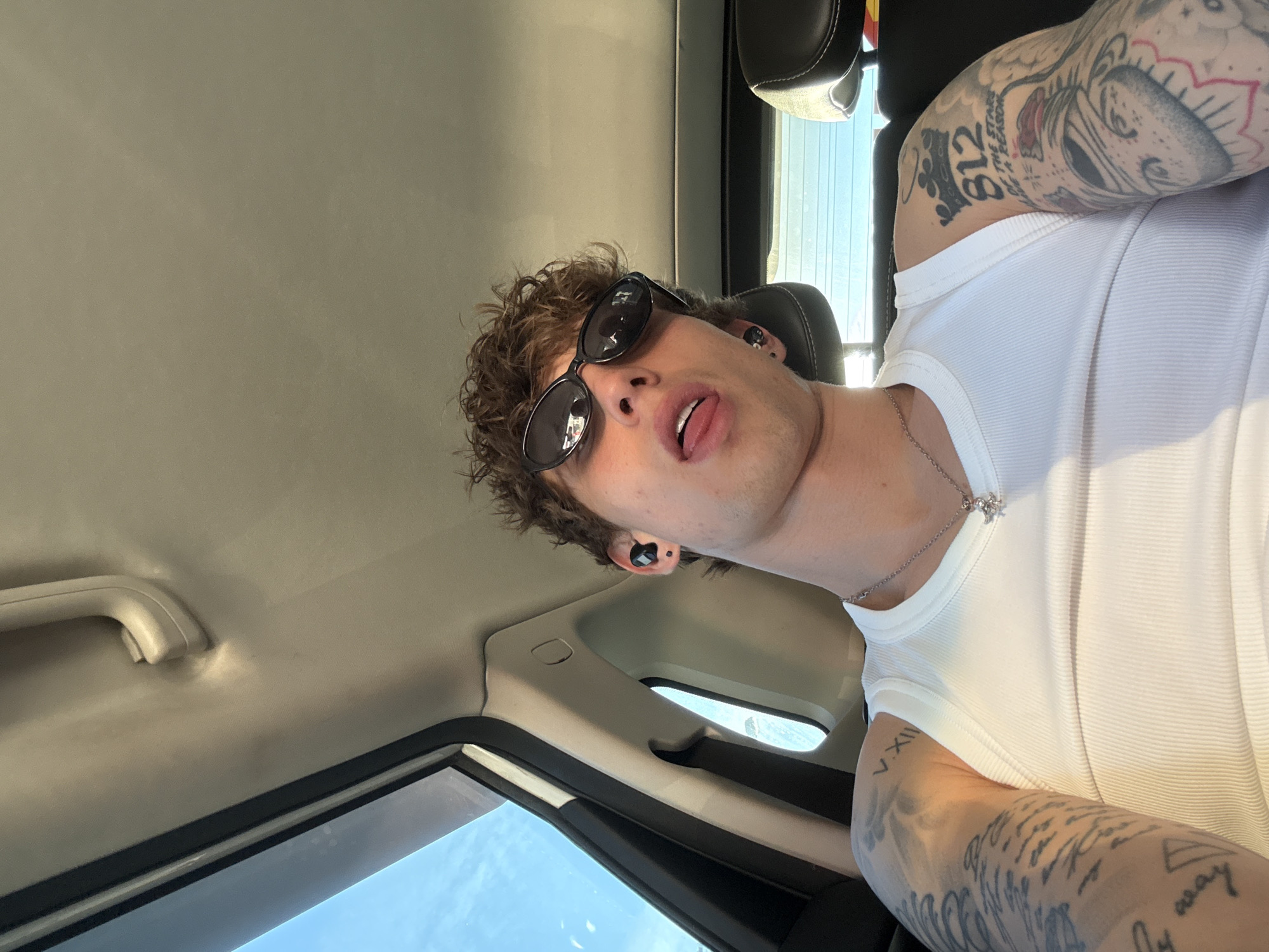 A man with curly brown hair and sunglasses sits in a car. He is wearing a white tank top and a silver necklace. His arms are heavily tattooed. The car's interior is visible, including the roof, window, and seat. The location is presumed to be in Khimki, Russia, based on the request, although the image itself contains no specific identifying markers of that location.