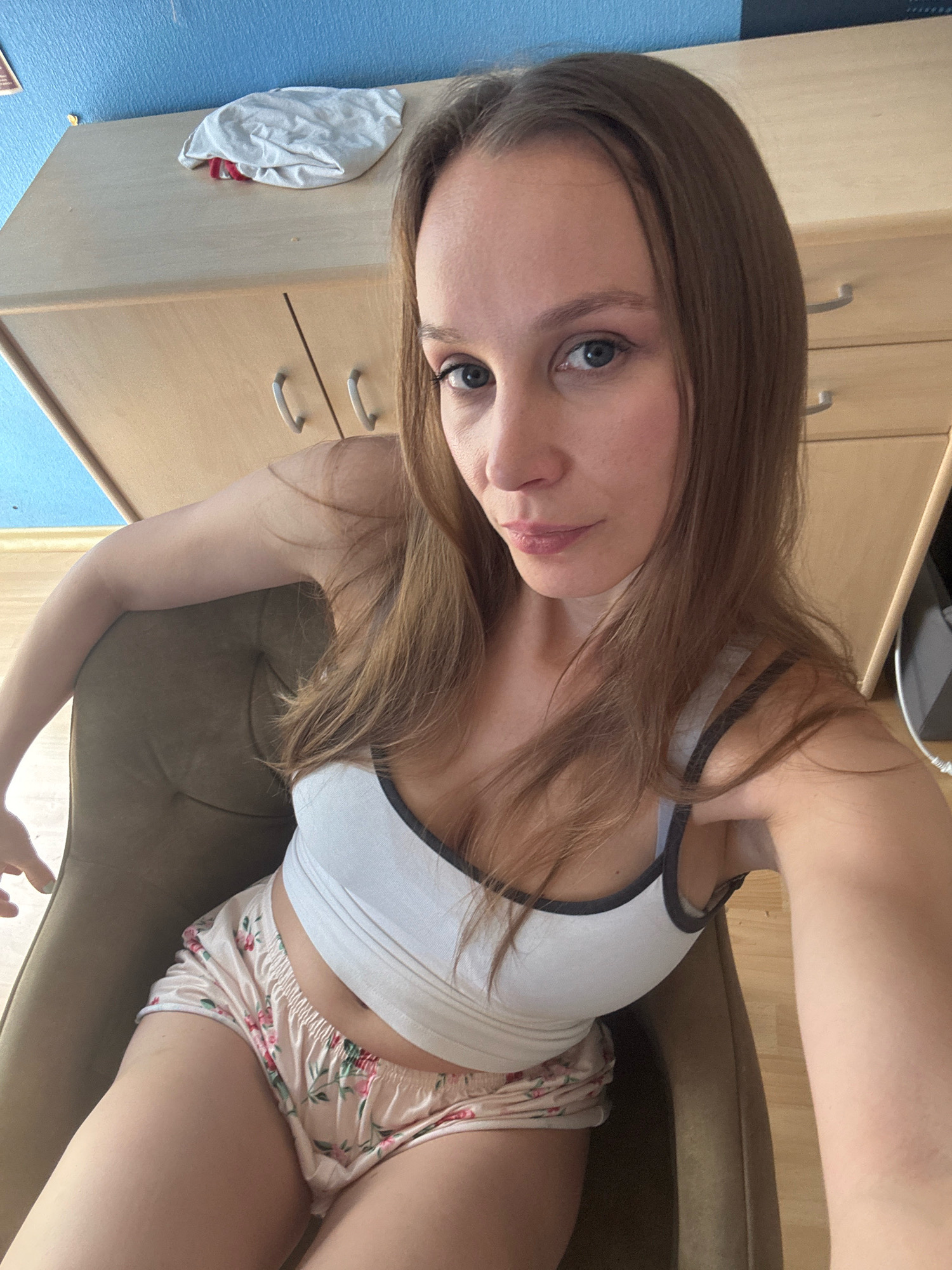 An individual is seated in a chair, facing the camera. The individual has long, brown hair and is wearing a white tank top with grey trim and floral patterned shorts. Behind the individual is a wooden dresser with two drawers, and a light blue wall. The scene is indoors.