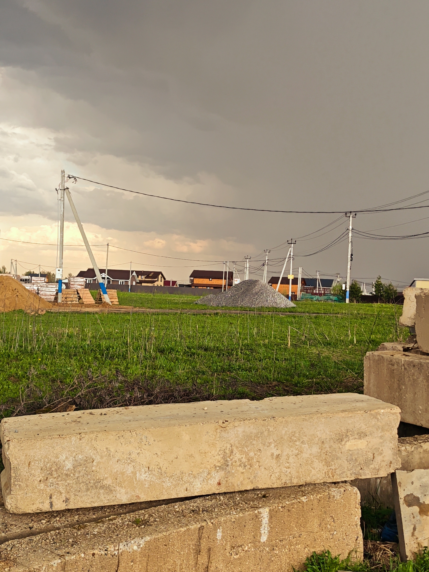 The scene depicts a rural or suburban setting, likely in Svistukha, Russia, based on the prompt. The sky is overcast with heavy, dark gray clouds. The foreground features a stack of large, rectangular concrete blocks, possibly construction materials. Beyond the blocks, there's a grassy field with some green vegetation. In the distance, there are residential houses, some with brown or orange roofs. Power lines and utility poles are visible, running across the landscape. There are also piles of materials, one appearing to be sand or gravel, and stacks of lumber. There are no people visible in the frame.