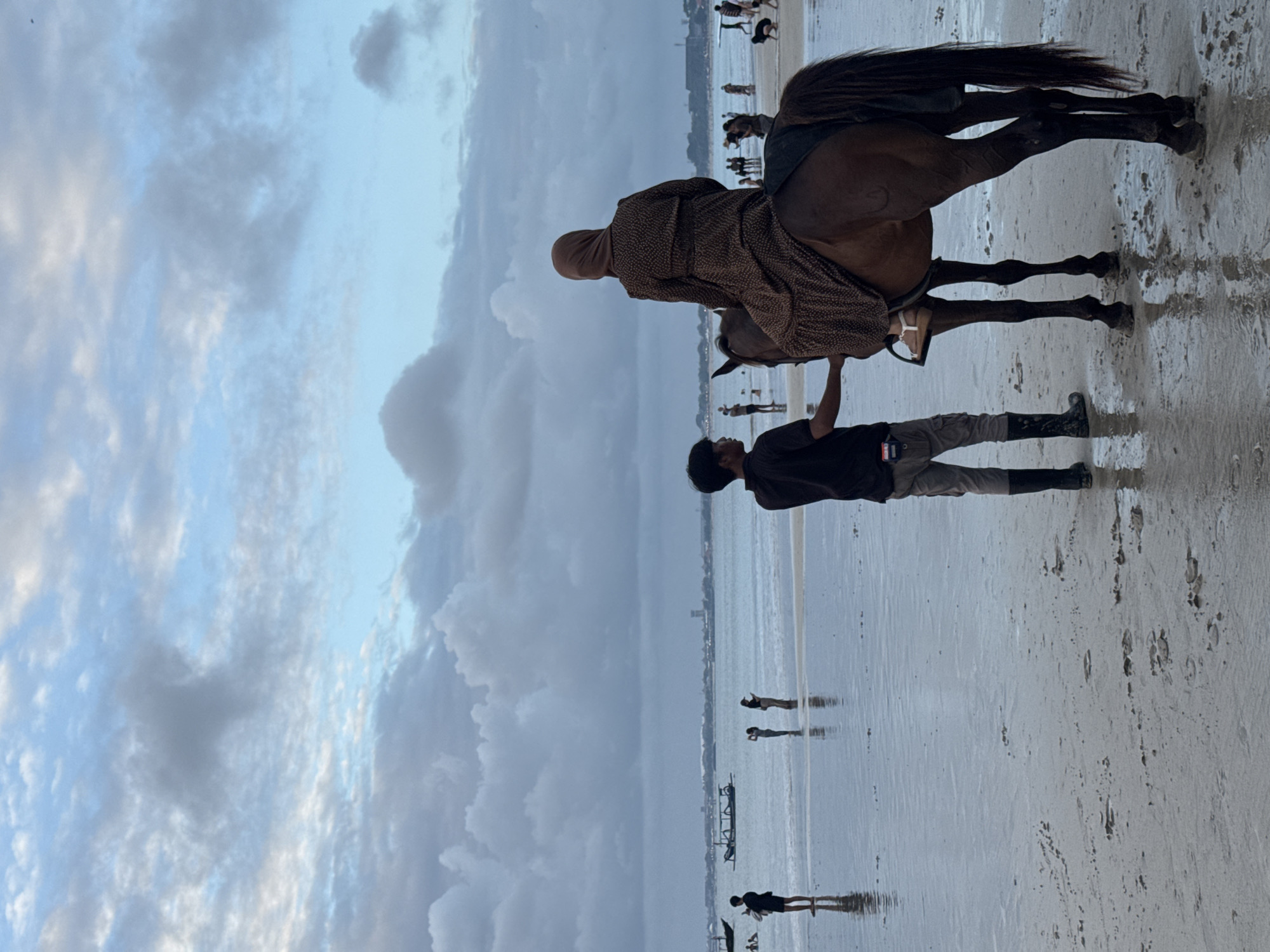 A woman wearing a brown patterned dress and a beige hijab is riding a brown horse on the beach. A man in a black shirt and gray pants is standing next to the horse, holding its reins. The beach is wet, with shallow water reflecting the sky and clouds. In the background, there are other people on the beach and in the water, as well as boats in the ocean. The sky is overcast with light blue and gray clouds. The location appears to be Kuta Selatan, Indonesia, possibly near a coastline with a backdrop of distant buildings.