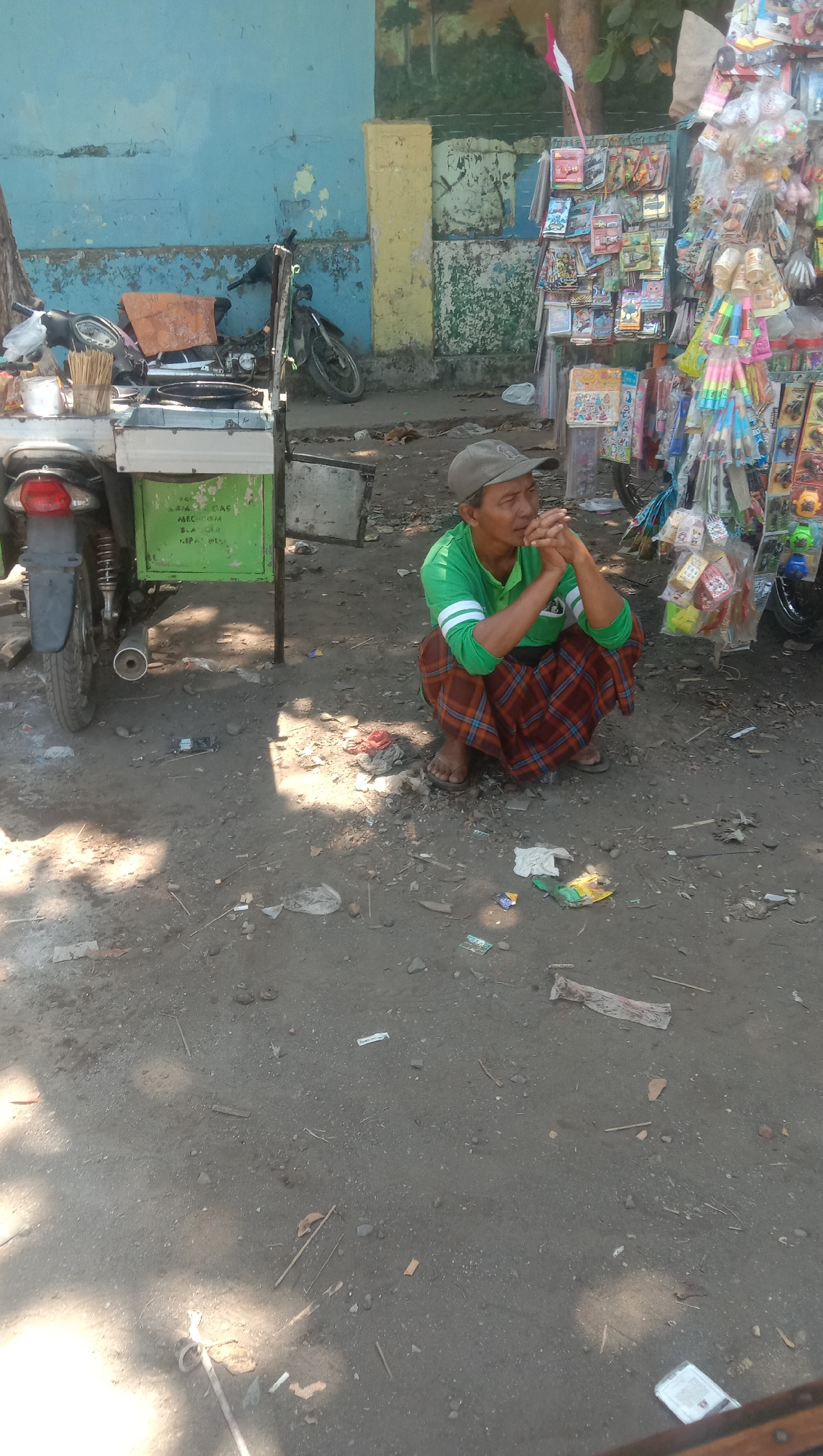 A man wearing a green long-sleeved shirt, brown and orange checkered sarong, and a grey cap is squatting next to a street stall selling small toys and novelty items. He is facing right, with his hands clasped in front of him, and appears to be looking off into the distance. The stall is colorful and filled with various plastic toys, keychains, and other small items. Behind the stall is a weathered wall painted blue and yellow, with some graffiti. To the left of the man, a motorcycle is parked next to a food cart. The ground is dirt and gravel, littered with small pieces of trash. The scene appears to be outdoors on a sunny day. The location is identified as Ngempit, Indonesia.