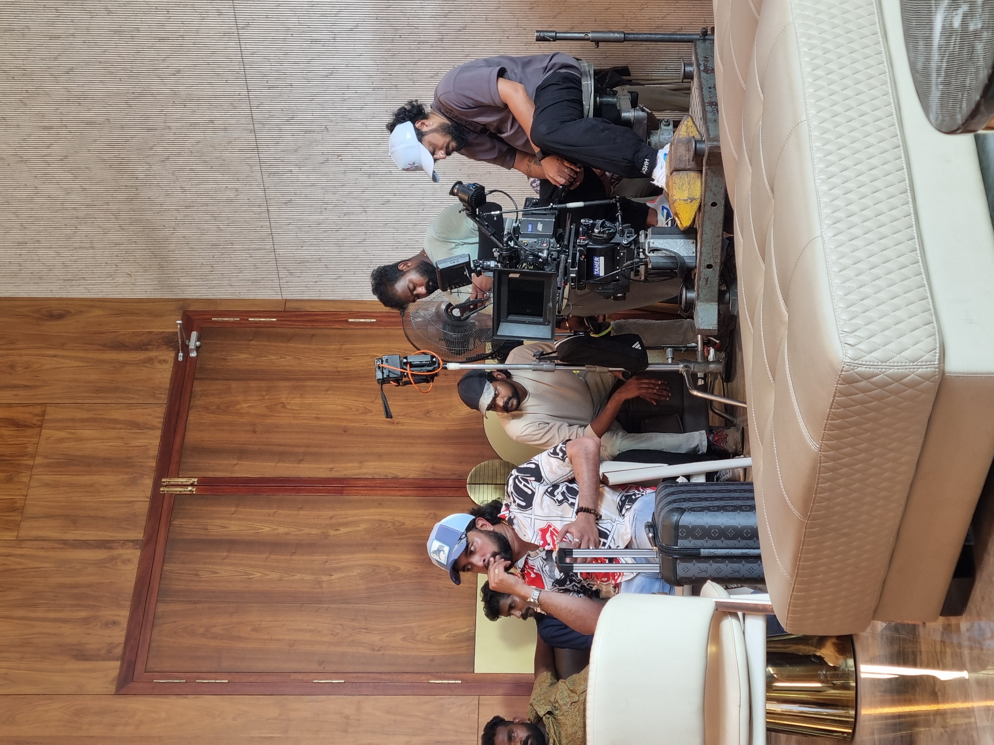 The image shows a scene inside a building in Kottukkara, India. Four men are visible, all appearing to be involved in a film or video shoot. One man in a grey shirt and white cap is leaning over a professional video camera mounted on a dolly, seemingly adjusting it. Another man in a green shirt is also working on the camera. Another man in a patterned shirt and blue cap is seated and looks like he's in discussion with others. A fourth man is sitting behind him. A large padded white sofa is next to the camera setup. The interior has wood-paneled walls and a horizontally striped ceiling. Suitcases are near the seated man in the patterned shirt.