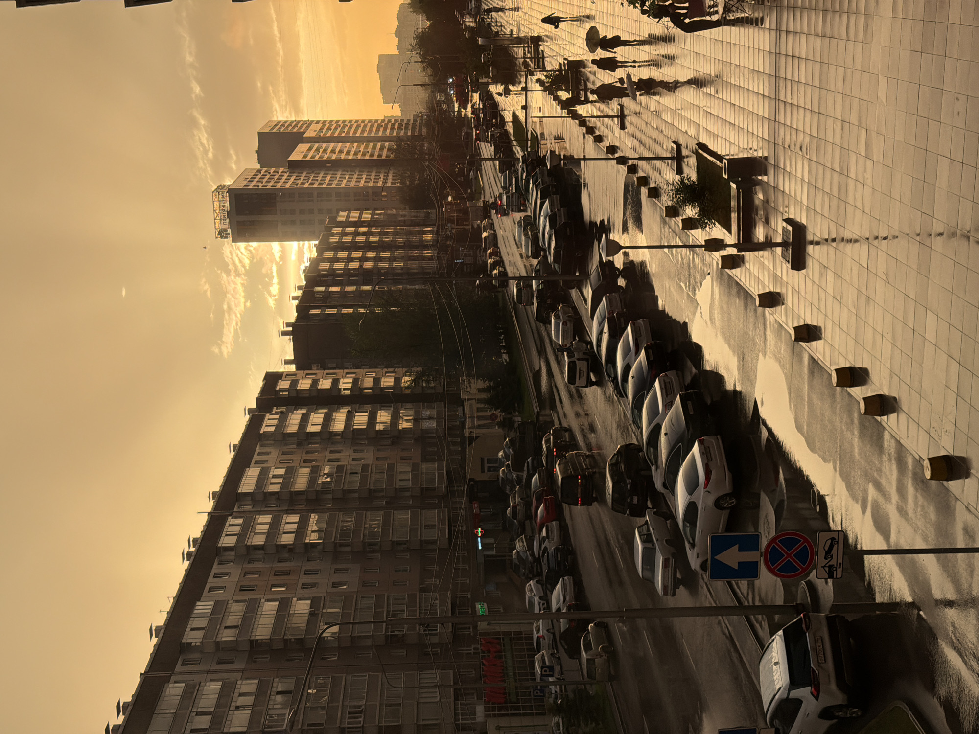 The image shows a city street in Krasnoyarsk, Russia, during a sunset. The sky is a warm, golden-orange, with wispy clouds. Tall apartment buildings line one side of the street, their windows reflecting the sunset. The other side of the street features a wet sidewalk, suggesting recent rain, with several people walking, some holding umbrellas. A row of parked cars is visible along the street. Streetlights and traffic lights are also present. The overall mood is serene, with the warm light of sunset casting long shadows. Visible text includes street signs and signs on buildings.