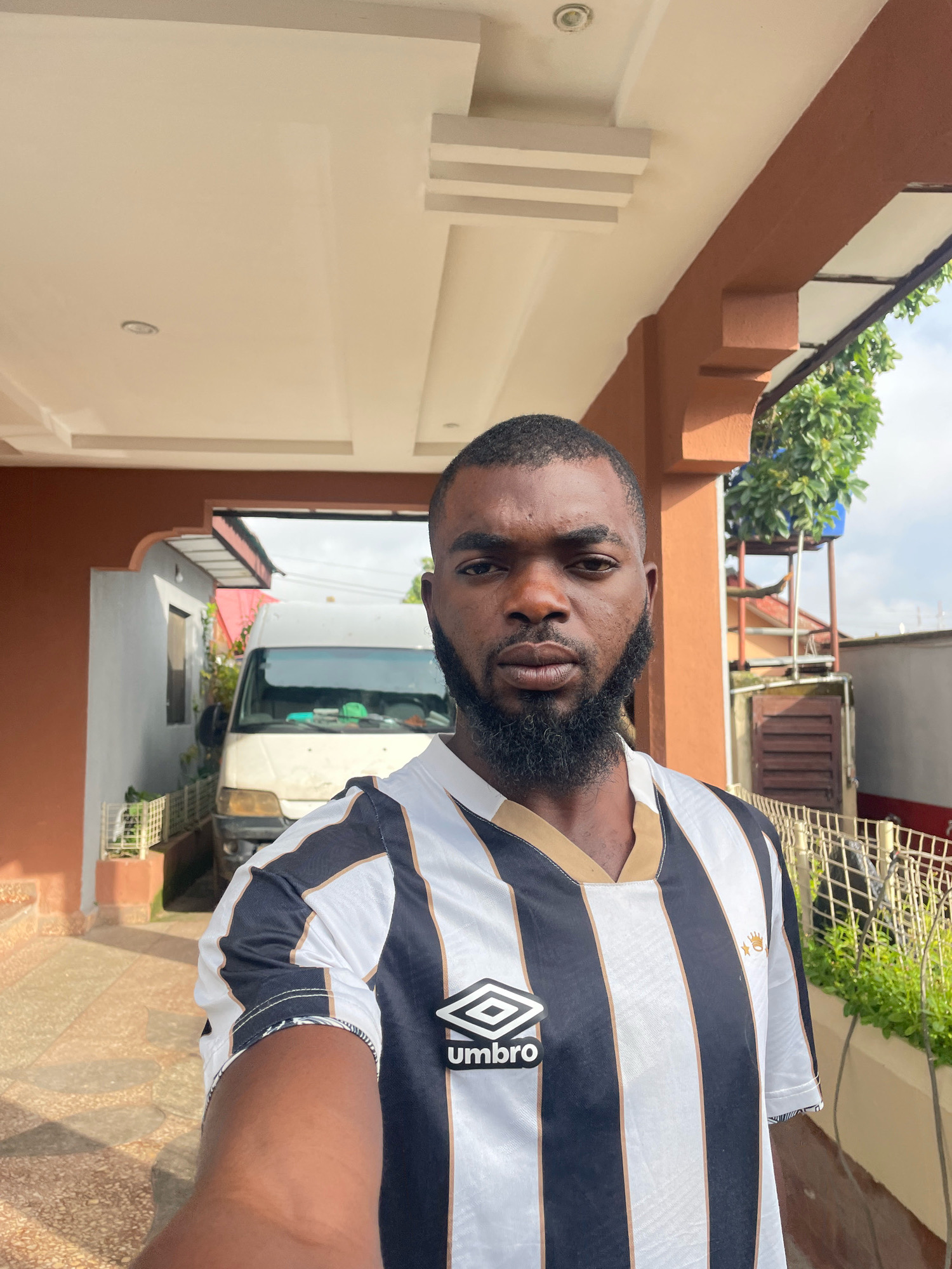 A man, identified as residing in Jos, Nigeria, poses for a selfie in front of a structure with a brown overhang. He is wearing a black and white striped jersey with gold trim and the Umbro logo. Behind him, a white van is parked under an overhang. The area appears to be a residential or commercial outdoor space, possibly a driveway or a courtyard, with a paved ground and some greenery visible. The lighting suggests it is daytime.