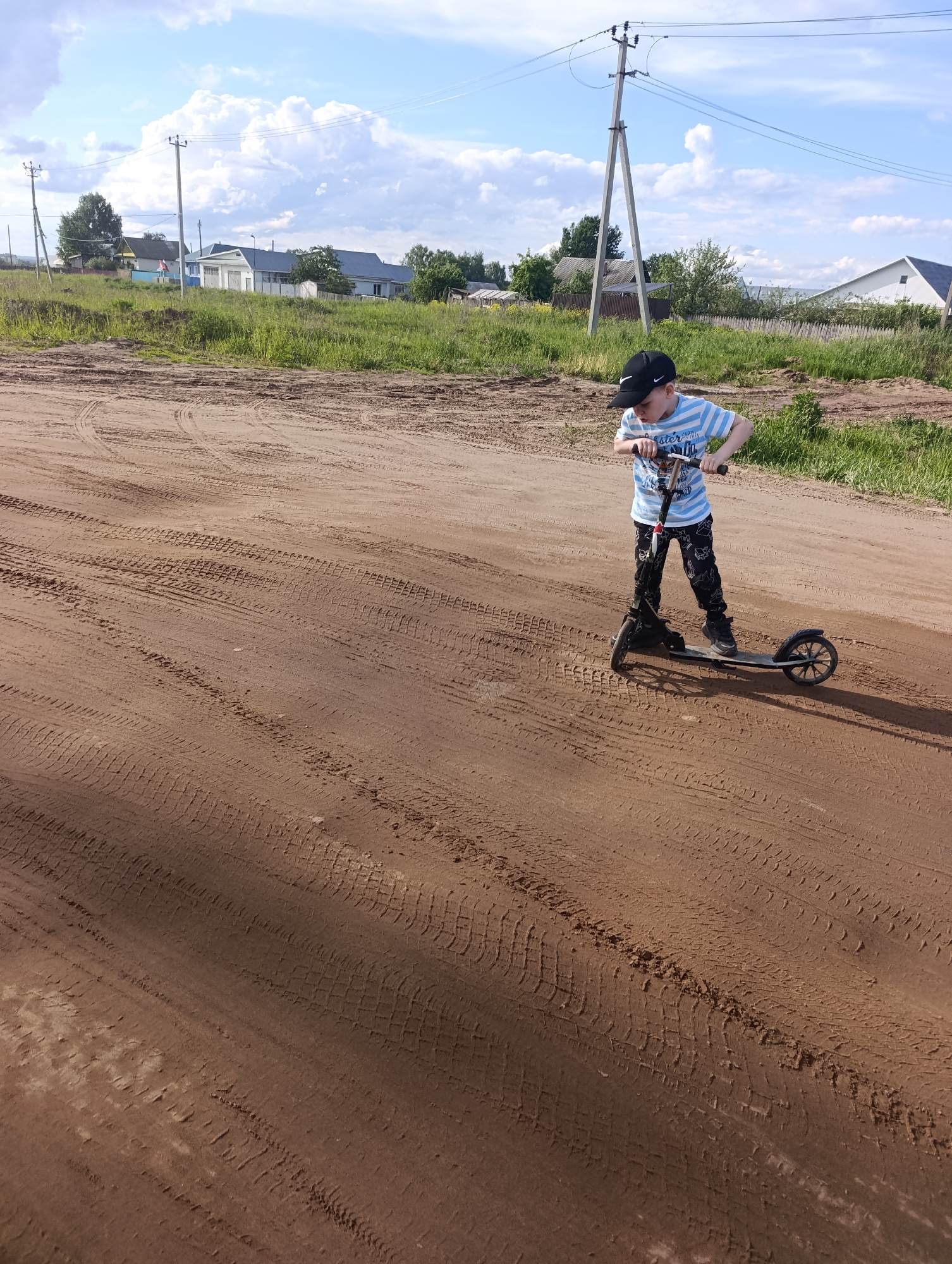 A young boy in a backwards baseball cap, striped shirt, and patterned pants rides a black scooter on a dirt road. The road surface shows extensive tire tracks. In the background, there is a grassy verge with a row of houses and utility poles. The sky is blue with scattered white clouds. The location is in the city of Chaikovsky, Russia.
