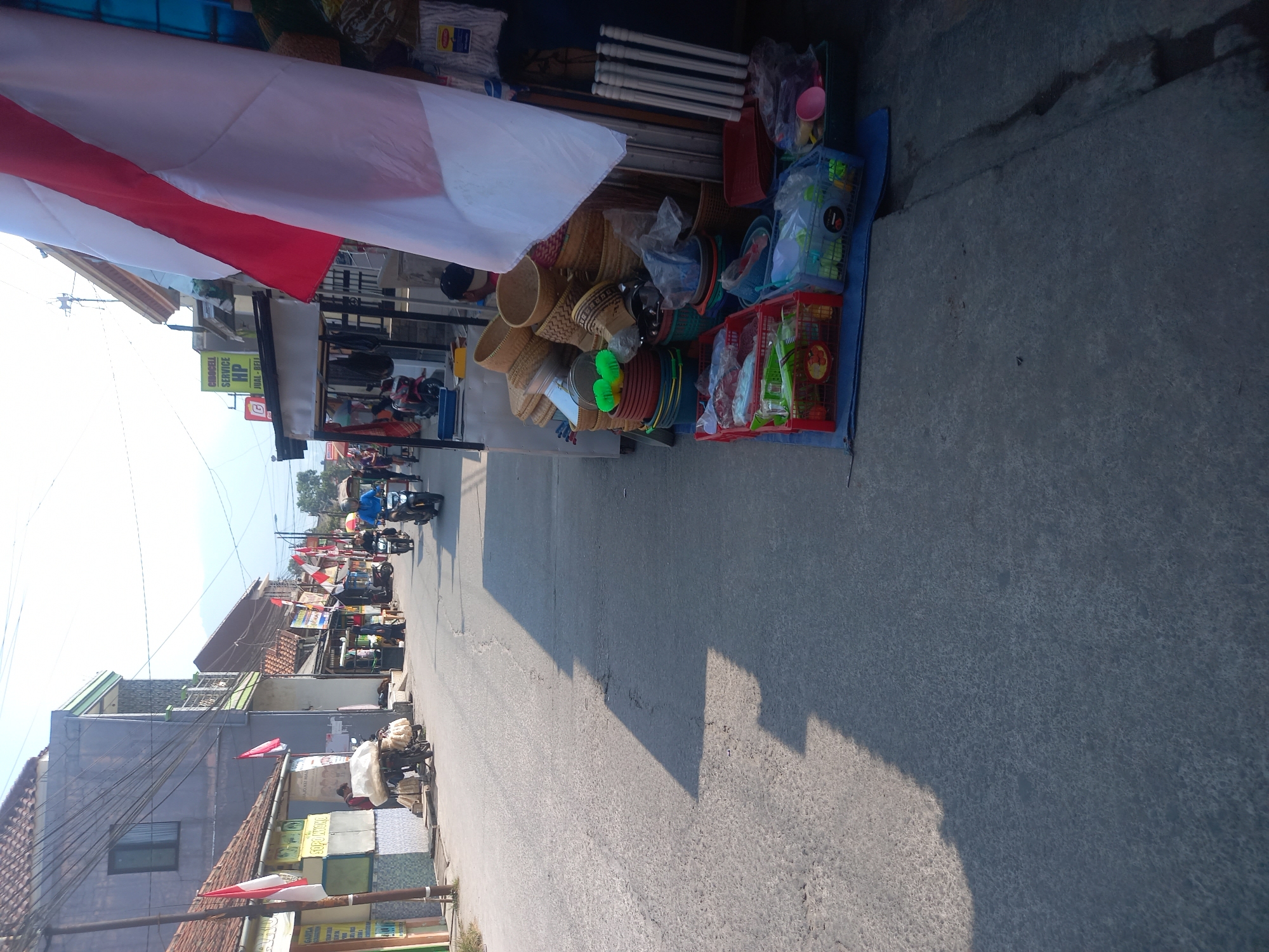 The image shows a street scene in Linggar, Indonesia, on a sunny day. The street is paved with asphalt and lined with buildings and market stalls. In the foreground, on the right side of the street, is a market stall displaying a variety of household items, including baskets, plastic containers, and cleaning supplies. A large Indonesian flag, red and white, is prominently displayed at the stall.
Further down the street, several people are seen. A man in a blue shirt is riding a motorcycle, and others are walking or standing near the shops. Some of the buildings appear to be shops or small businesses, with signs in Indonesian. There are also several parked motorcycles and a cart on the side of the road. The overall impression is of a bustling local street with active commerce. The environment appears to be a typical street in a residential or commercial area in Indonesia.
