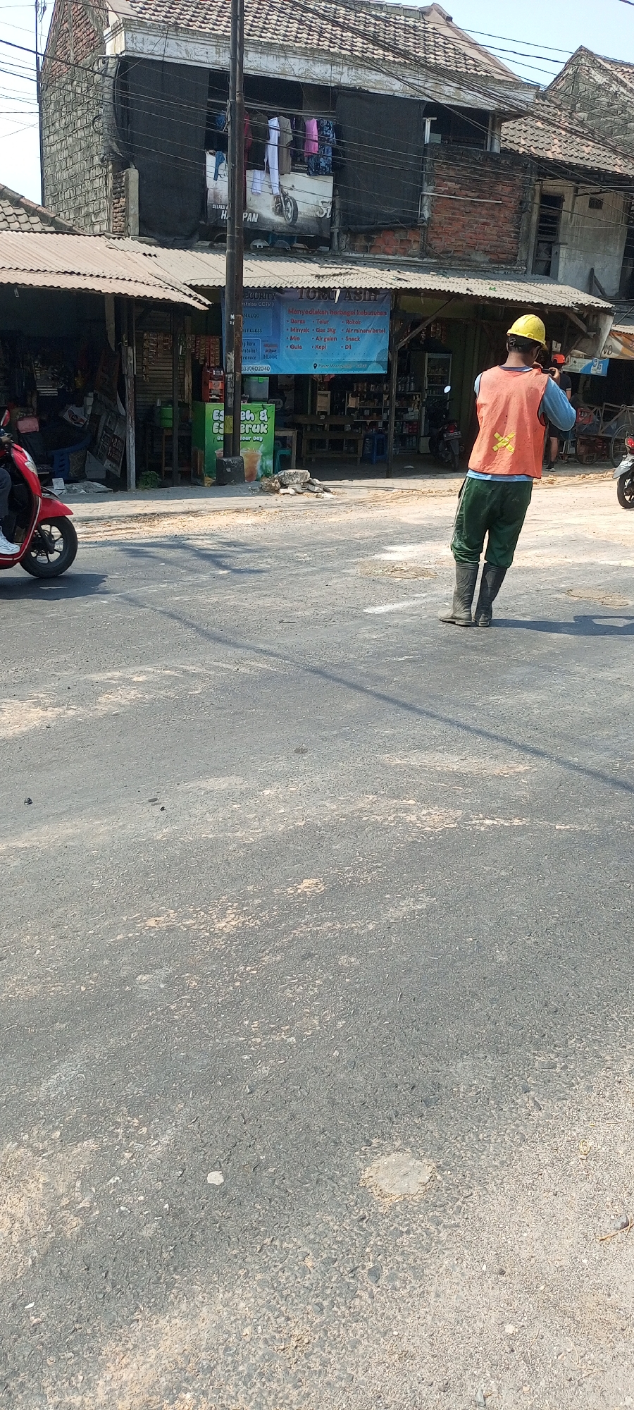 In Moropelang, Indonesia, an individual wearing a yellow hard hat and an orange reflective vest is standing in a street, facing away from the camera. They are also wearing green pants and dark boots. Behind them, there is a multi-story building with a tiled roof and what appears to be laundry hanging from an upper level. In front of the building is a covered market or shop area with various items displayed. To the left of the frame, a red scooter is partially visible, with its rider also mostly out of view. The street is paved with asphalt and shows signs of wear. The lighting suggests it is daytime.