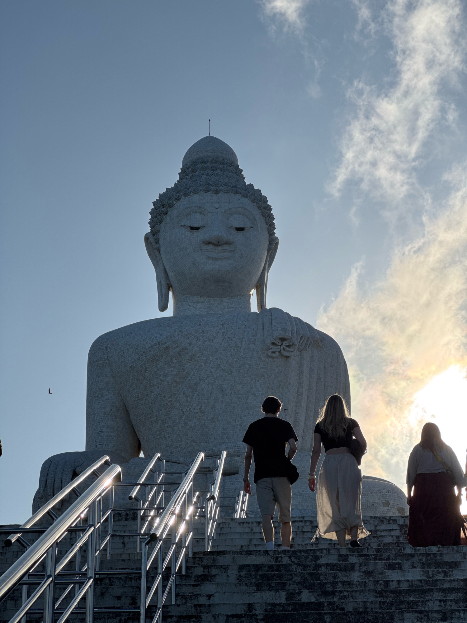 This image depicts the Big Buddha statue in Karon, Thailand. Three individuals, silhouetted against the bright sky and a bird in flight, ascend a wide concrete staircase. The staircase features gleaming metal handrails that reflect the sunlight. The colossal white statue of the Buddha dominates the background, with its serene face looking downwards. The sky is a clear blue with scattered, wispy clouds, and the sun is visible, casting a warm glow on the scene.