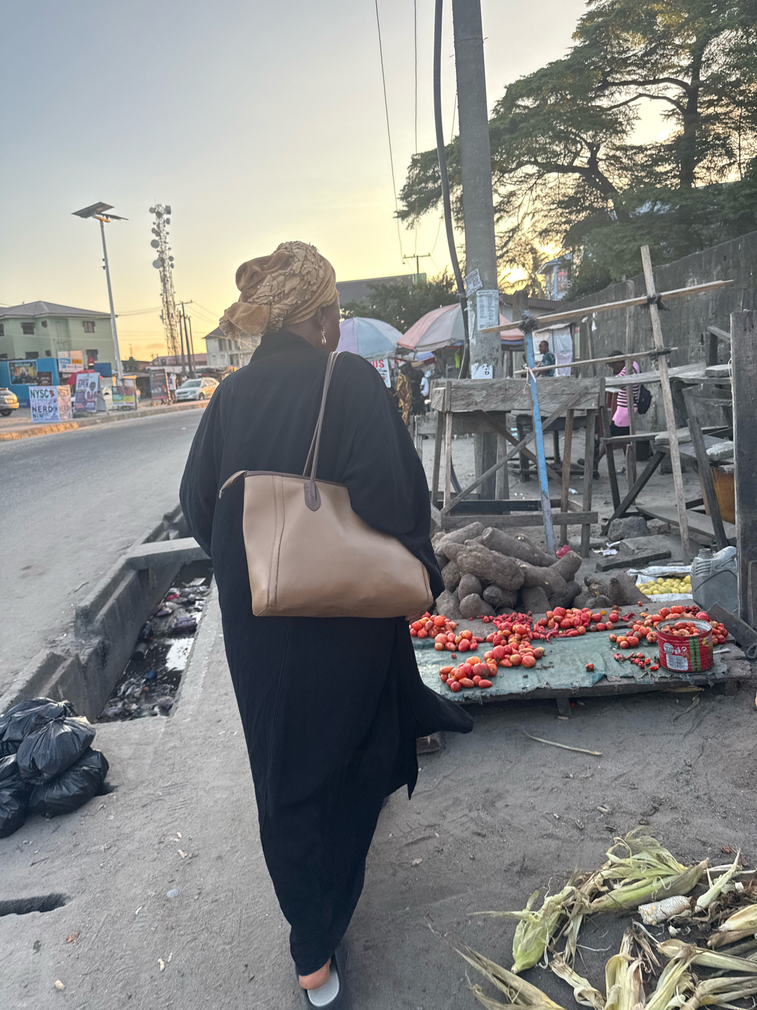 The image depicts a street scene in Ado, Nigeria, at dusk. A woman, viewed from behind, walks away from the camera on a concrete path. She wears a black abaya and a gold headscarf, and carries a tan leather shoulder bag. To her left is a drainage ditch filled with refuse. To her right, a roadside market is set up. Stalls, made of rough wooden structures and covered with tarps, display piles of yams and tomatoes. A few individuals are visible in the background, tending to the market or walking along the street. The street is paved, and in the distance, vehicles are on the road, suggesting ongoing urban activity. A cell phone tower and utility poles are also visible against the hazy twilight sky. The ground is a mix of dirt and concrete, with discarded corn husks scattered in the foreground.