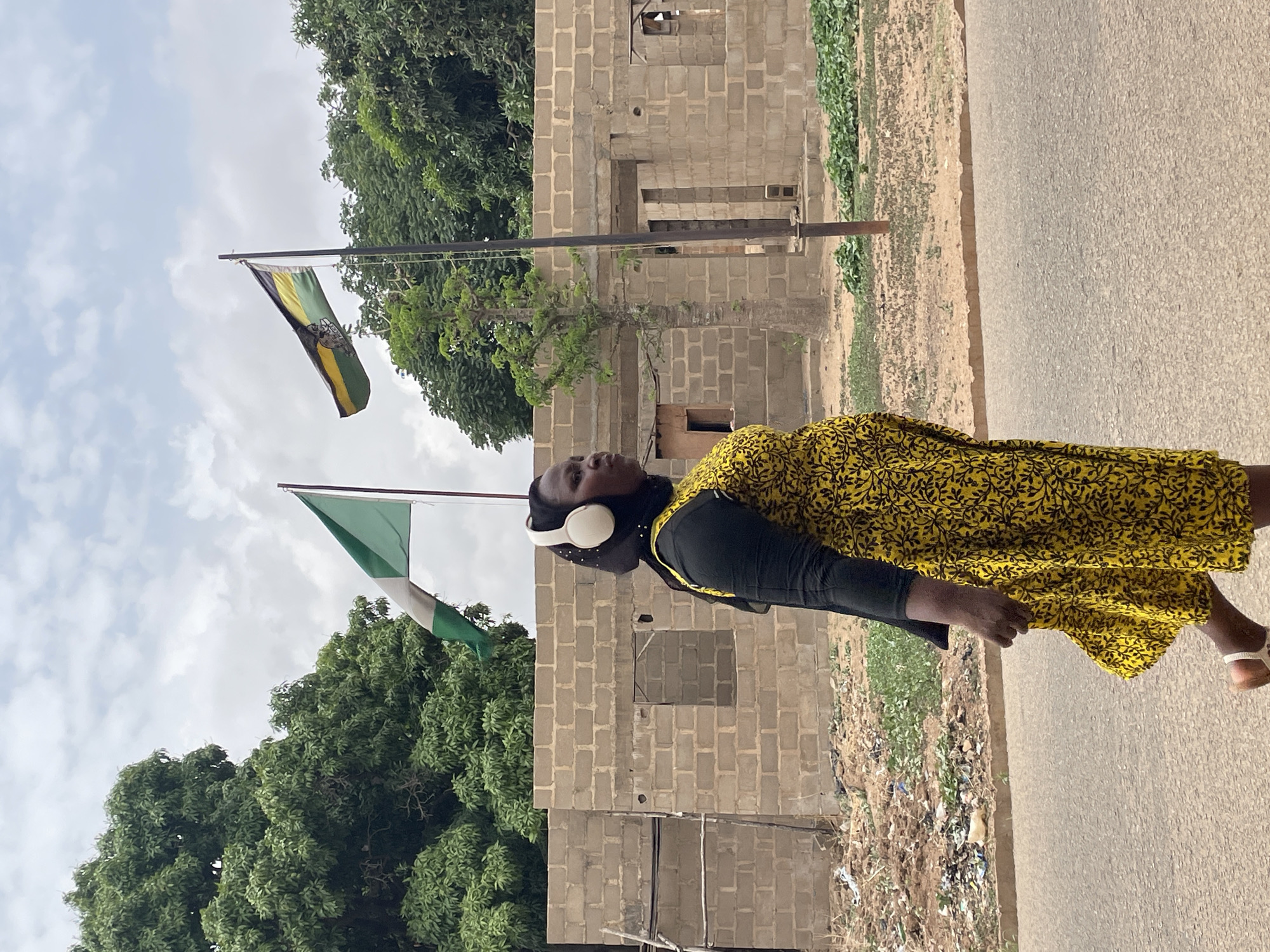 A woman is walking on a road in Jos, Nigeria. She is wearing a yellow dress with black patterns, a black hijab, and white sandals. She also has white headphones on. A partially constructed building is in the background along with two flags, one green and white, and the other green, yellow, and black. The environment consists of trees, a road, and debris on the side of the road. The sky is overcast.