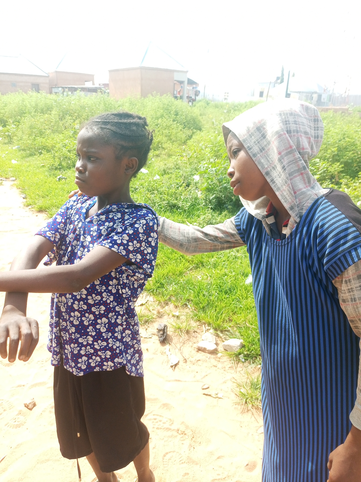 Two young children, one on the left and one on the right, stand in a dusty, outdoor environment. The child on the left is wearing a blue short-sleeved shirt with a white floral pattern and black shorts. They have their arms crossed and are looking to the left. The child on the right is wearing a blue and black striped long-sleeved shirt with a checkered hooded jacket over it, the hood pulled up. They are reaching out towards the child on the left. The background consists of green vegetation and some buildings in the distance, suggesting a semi-urban or rural setting. The ground is sandy and appears to be a path or unpaved area. There is no explicit indication of the city of Tempure, Nigeria within the image itself.
