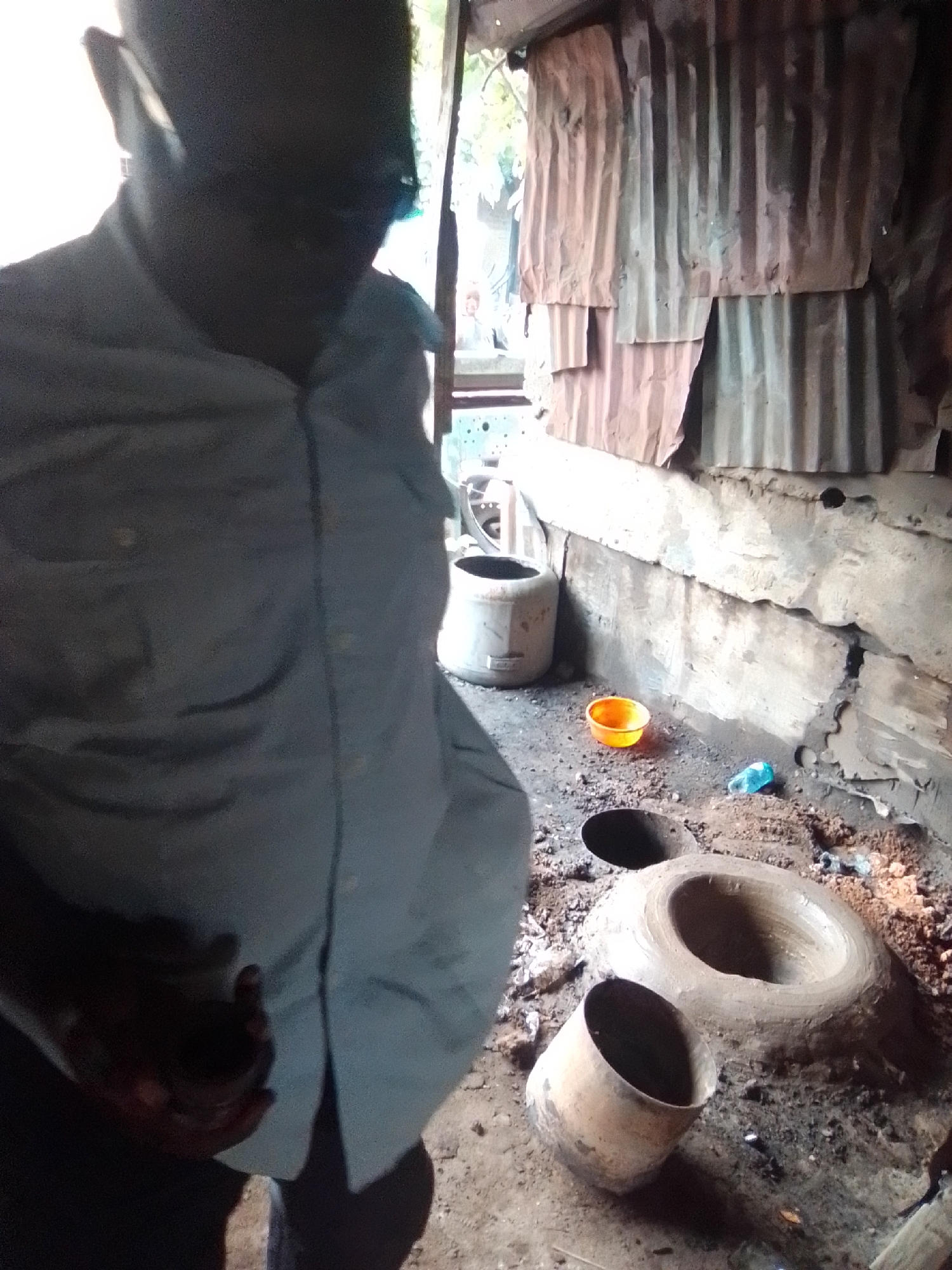 The image depicts a person in the foreground, partially obscured by shadow, wearing a light-colored collared shirt. The background reveals a rustic, open-air structure constructed from corrugated metal sheets and rough wooden planks. The ground is covered in dirt, ash, and debris. Several clay pots of various sizes are present, some appearing to be used for cooking or as part of a kiln setup. A small orange bowl and a blue plastic bottle are also visible on the ground. The scene suggests an outdoor workshop or cooking area, likely in a rural or informal settlement. The setting is consistent with an area in Arawa, Nigeria, given the materials and overall aesthetic of the environment.