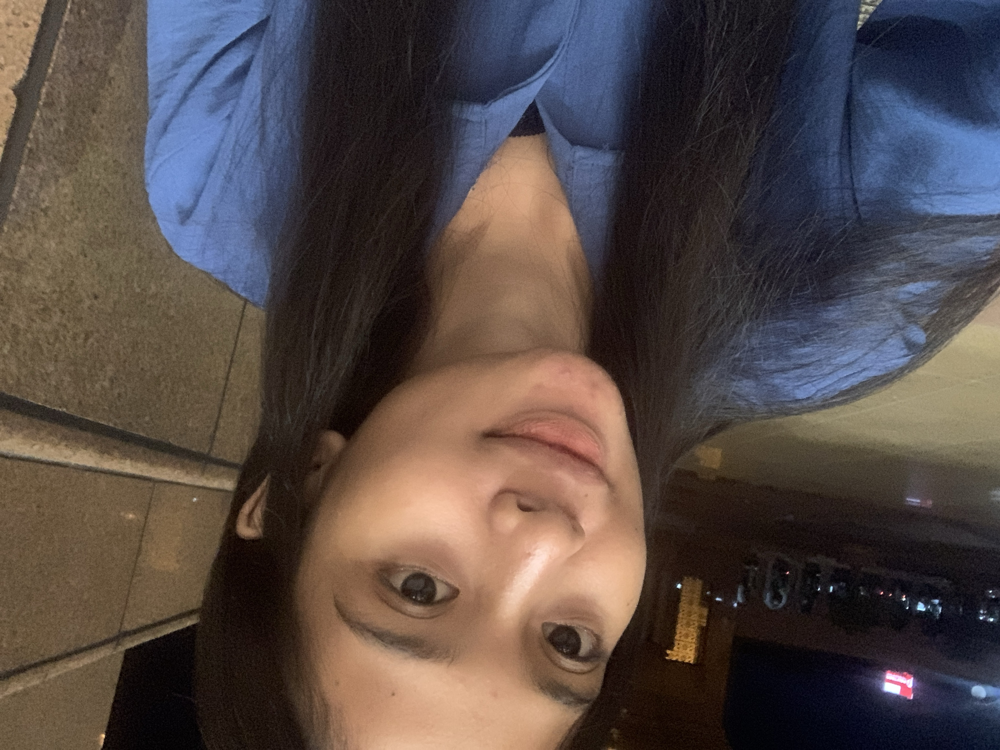 The image is a close-up selfie of a young woman, captured in a low-light setting. The photograph is oriented vertically, showing the woman's head and shoulders as she looks directly at the camera with a neutral to slightly smiling expression. Her fair-skinned face is relatively well-lit, highlighting her dark eyes and long, dark hair that frames her face. She is wearing a blue collared top or blouse.
To her left, the background consists of a textured, light brown or grey surface, possibly a stone wall or ground. To her right, the scene recedes into a dark, out-of-focus area punctuated by blurred urban lights, including red and white glows, which are characteristic of an outdoor, night-time environment in a city. This suggests the photo was taken outdoors in the evening or at night. The location context provided indicates this scene is in Samarinda, Indonesia. No discernible text is visible within the image.