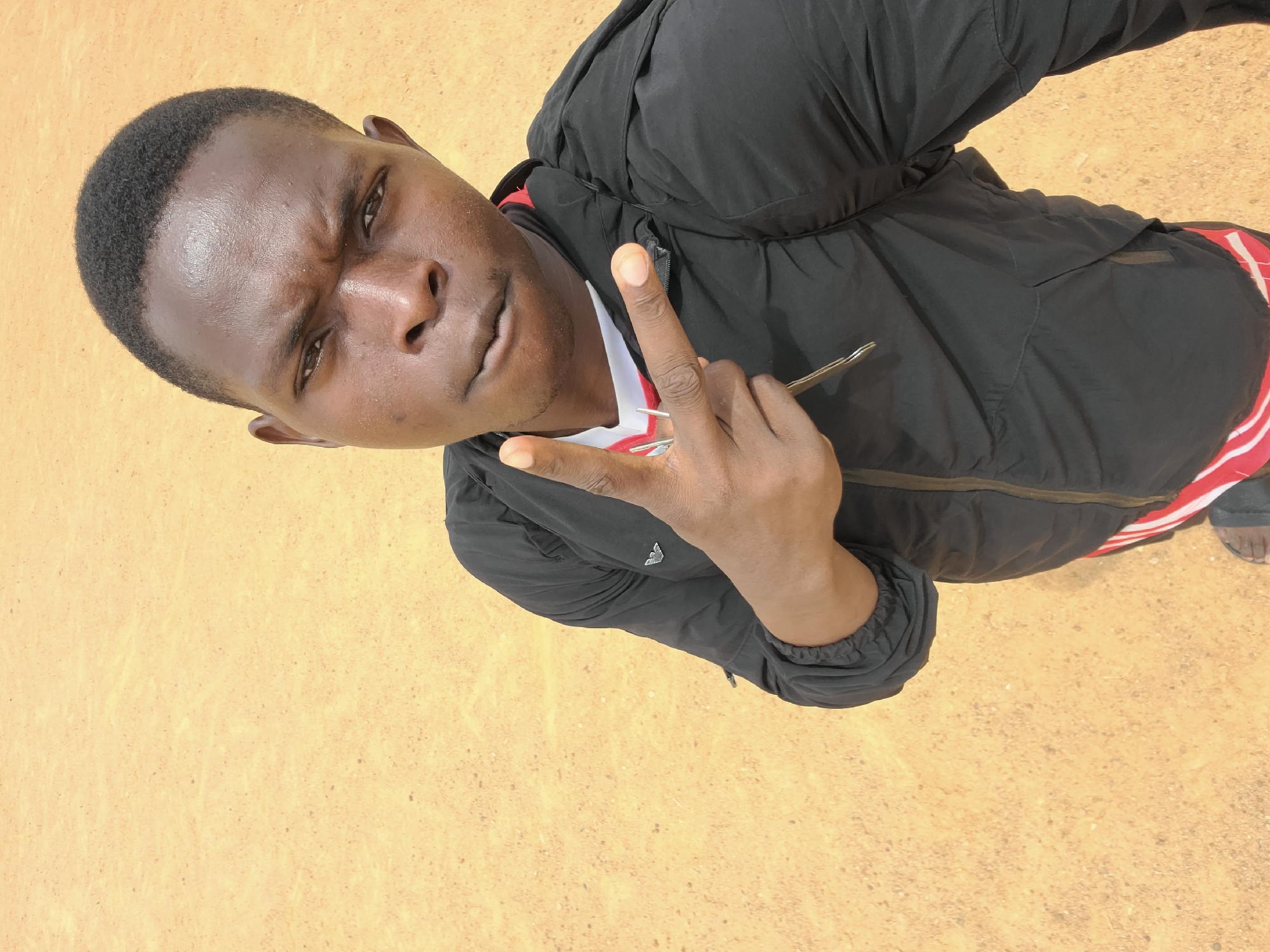 A medium-shot, high-angle photograph shows a Black male holding a metal object between the fingers of his left hand. He is wearing a black jacket, a white and red top underneath, and appears to be outdoors. The ground is a light brown or tan color. The location is assumed to be in the city of Bukuru, Nigeria.