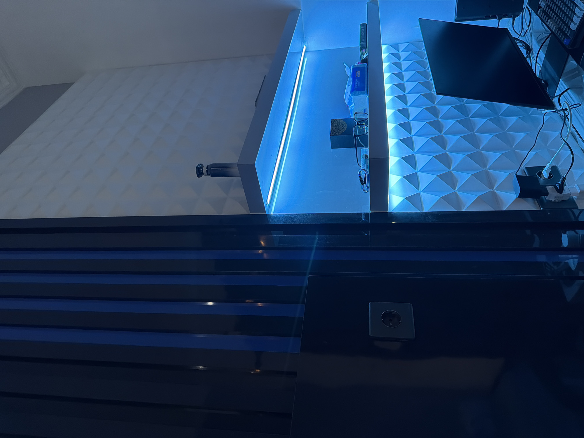 A dimly lit room, possibly an office or study, features a modern design with dark blue and white elements, enhanced by vibrant blue LED lighting. On the left, a dark blue panel with vertical stripes, some in a brighter blue, occupies the foreground. A single electrical outlet is visible on this panel. To the right, white geometric wall panels create a textured background. Two shelves are illuminated by blue LEDs. The upper shelf holds a dark cylindrical object and a remote control. The lower shelf contains a small dark box, a pair of glasses, a pack of tissues, and other small items. In the lower right, a black computer monitor and keyboard are visible on a desk, with cables connecting to various devices. The overall impression is one of a contemporary, technologically equipped space with ambient lighting.