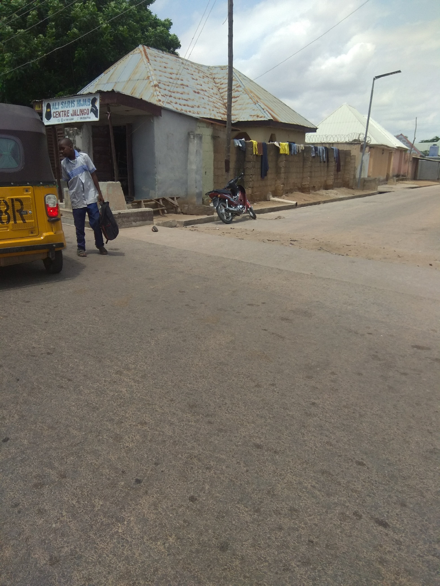 This is a medium-distance shot of a street scene in Jalingo, Nigeria, featuring a yellow auto-rickshaw on the left, a man walking with a bag, and buildings on the right. The auto-rickshaw, with a license plate partially visible, is parked on the side of the road. A man wearing a light-colored shirt and dark pants is walking away from the rickshaw, carrying a dark bag over his shoulder.
Behind the man and the rickshaw, there is a building with a corrugated metal roof and a sign above the entrance that reads "ALI SARIS HIJAB CENTRE JALINGO". The building appears to be a shop or a business establishment. Further down the street, there are other buildings with various roof types, including a white pitched roof with barbed wire fencing in front of it. Clothes are hanging on a line strung between two posts in front of one of the buildings. A red motorcycle is parked on the side of the road near the buildings. The street is paved with asphalt, and there are some patches of bare earth and debris. The sky is cloudy, with patches of blue visible. The overall impression is that of a typical urban street in a Nigerian city.
