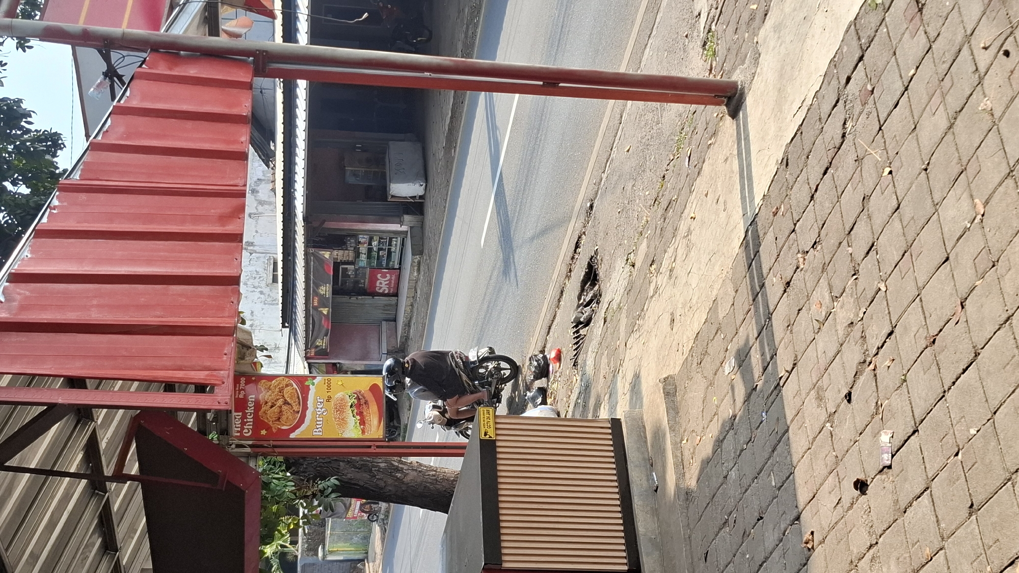 This is a medium-shot, eye-level, natural-light photograph of a street scene in Cilaku, Indonesia. In the foreground, a paved sidewalk made of rectangular bricks runs diagonally from the bottom left to the middle right of the frame. A beige and black modular kiosk-like structure is on the far left of the sidewalk. A few fallen leaves and debris are scattered across the bricks, with a small pile of currency visible near the bottom left corner.
The middle ground shows a paved road with a white line marking the center. A motorcycle, viewed from the side, is parked on the shoulder of the road. Two individuals wearing dark helmets are seated on the motorcycle. The driver is wearing a dark long-sleeved shirt and dark shorts, while the passenger, behind the driver, is wearing a dark long-sleeved shirt and dark pants. Two black plastic bags are on the ground next to the motorcycle.
In the background, on the left side of the road, a tree stands next to a red and yellow advertisement sign for "Fried Chicken" and "Burger." Behind the sign, a row of buildings forms a marketplace or commercial strip. One building has a prominent red corrugated metal awning. Another building, further down the street, has a sign that reads "SRC." The overall environment suggests a daytime scene in an urban or semi-urban area with commercial activity.