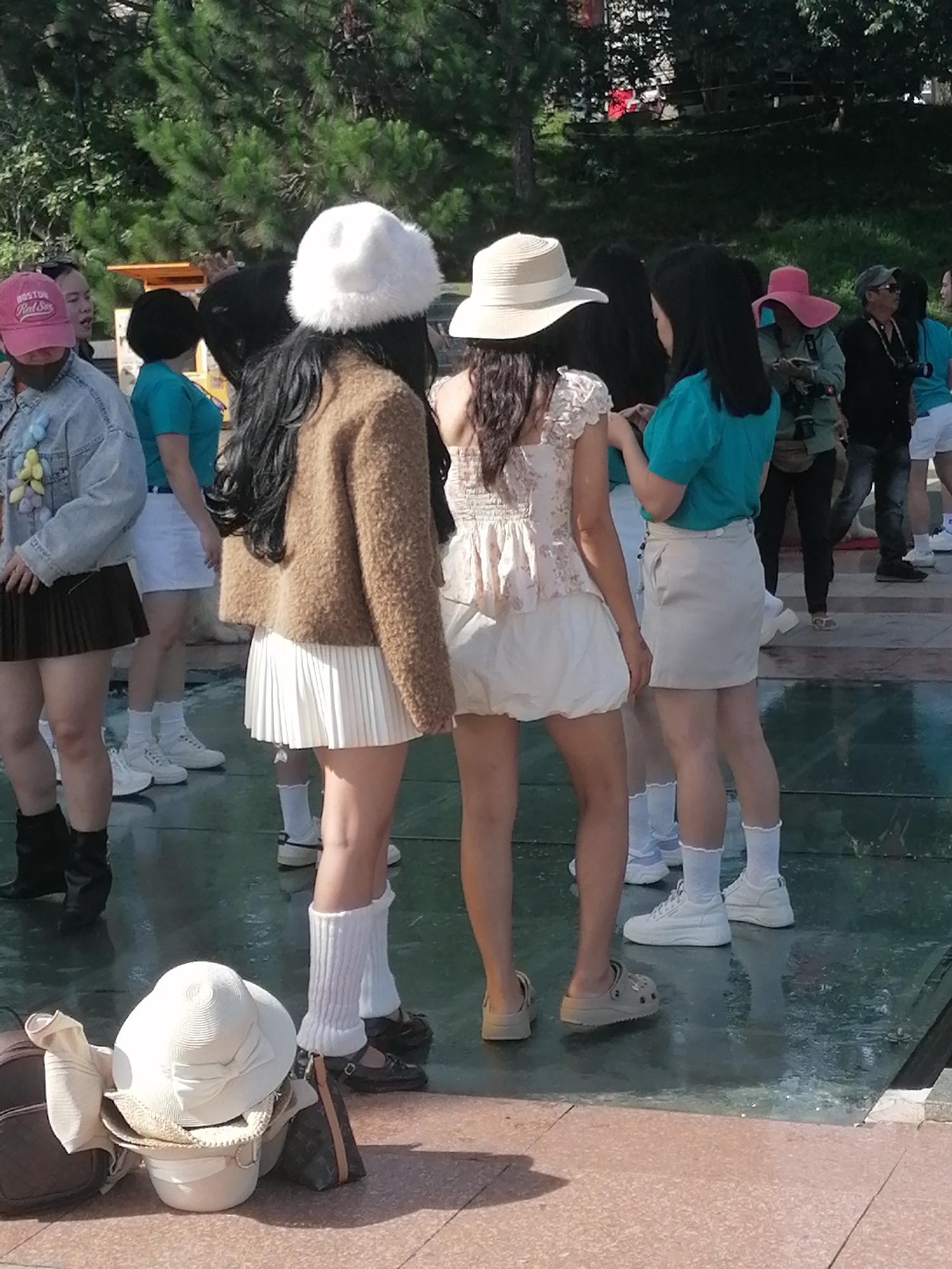 The image shows a group of women standing on a wet, reflective surface, possibly a plaza or pathway, in Da Lat, Vietnam. The women are dressed in casual, summery attire, with several wearing hats. In the foreground, to the left, are two white hats and two handbags on the ground. In the background, trees and foliage are visible, suggesting an outdoor setting. The overall impression is of a relaxed, informal gathering.